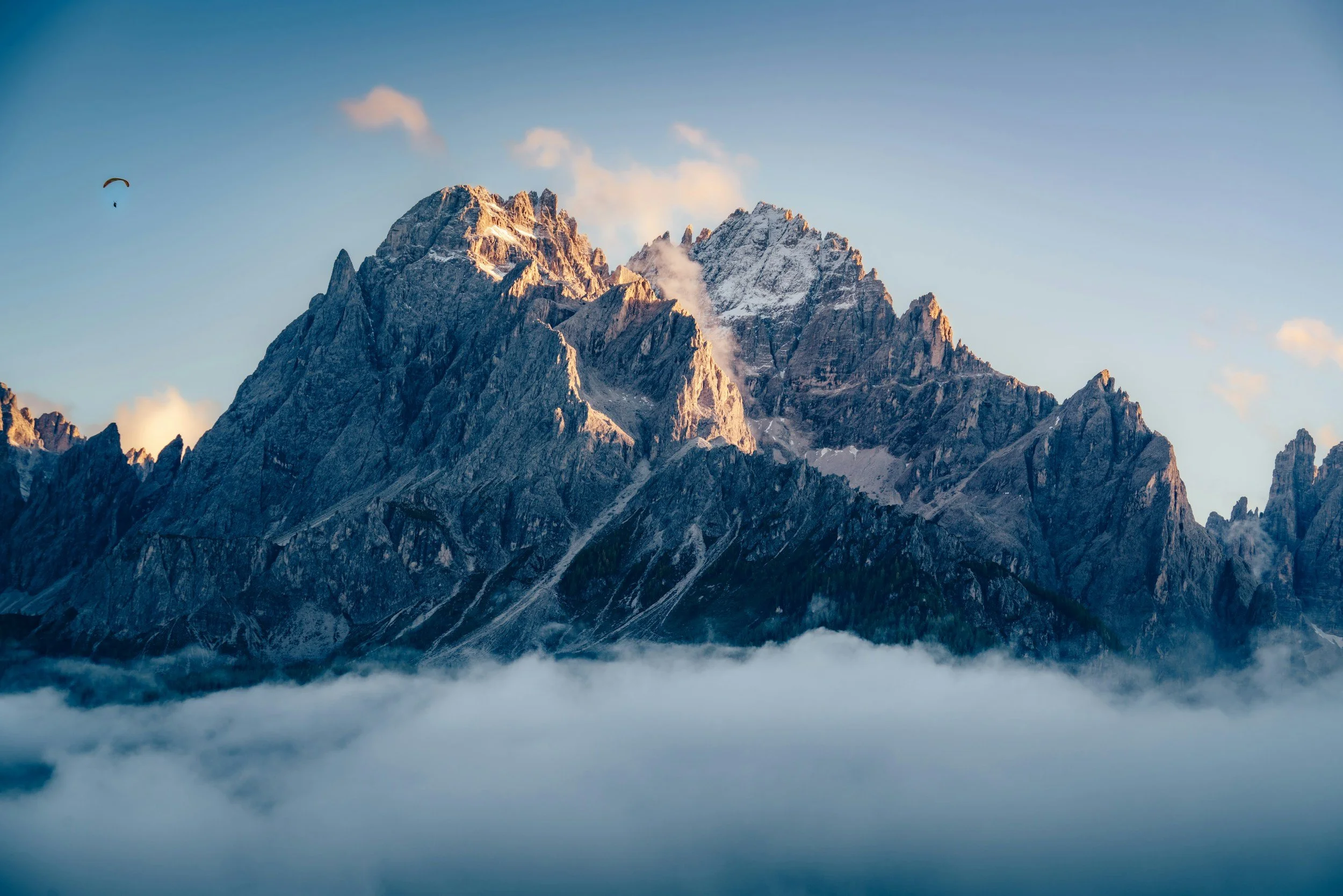 Snow-capped mountain peaks in the distance with clouds and fog in the foreground, clear blue sky above, and a paraglider in the distance.