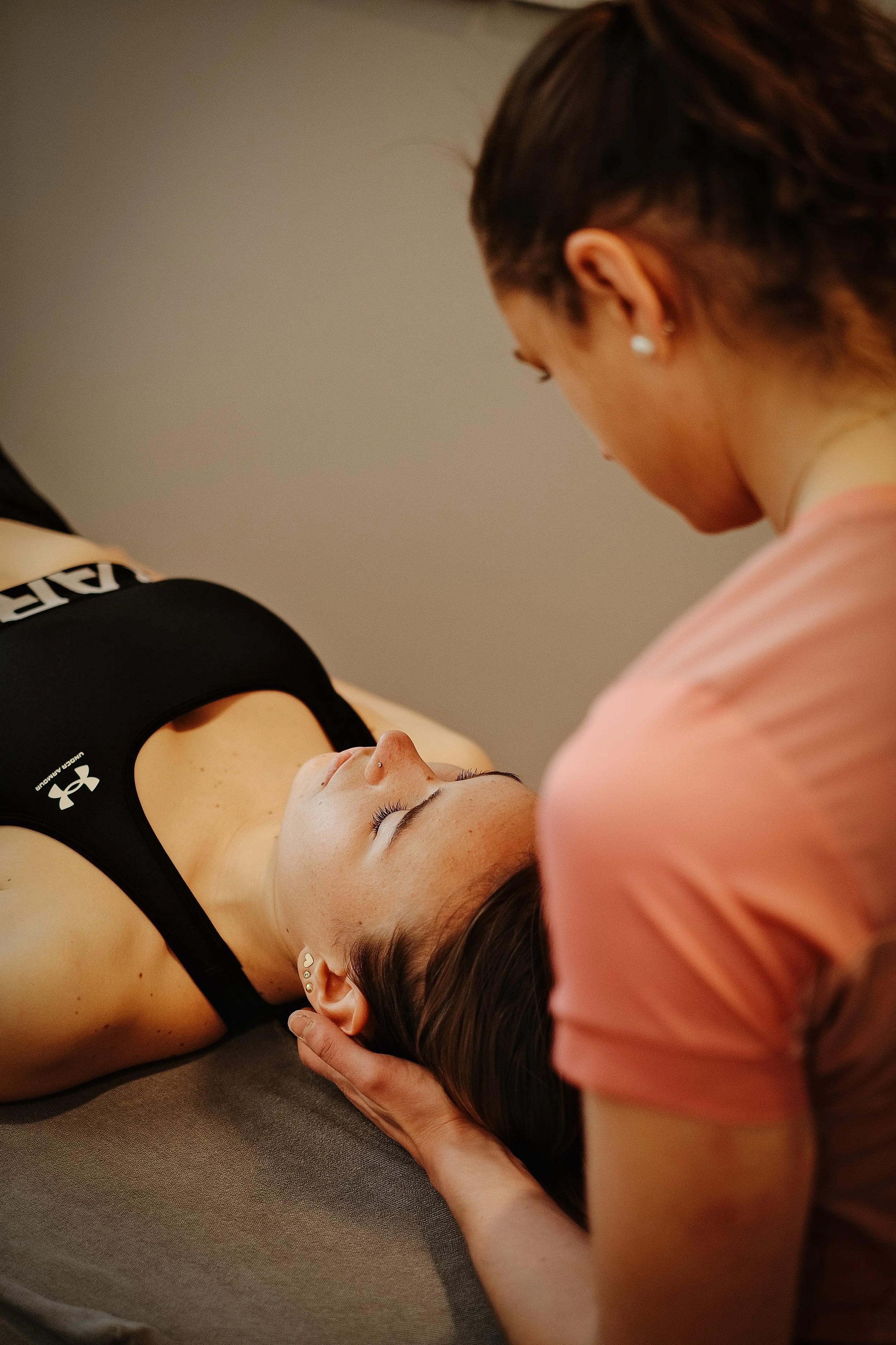 A woman with long dark hair lying down in a sports bra, receiving a head massage from another woman with short dark hair and earrings.