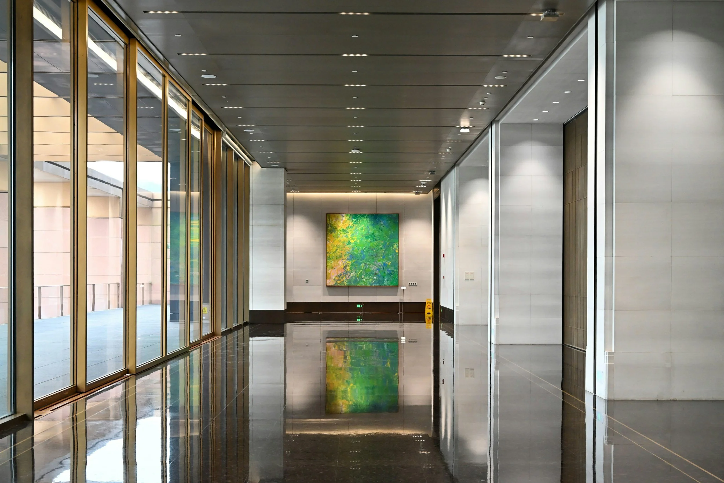 Modern interior hallway with glass walls on the left, sleek ceiling with small lights, and a large colorful abstract painting on the back wall. Reflective black tiled floor with a yellow caution sign near the wall.