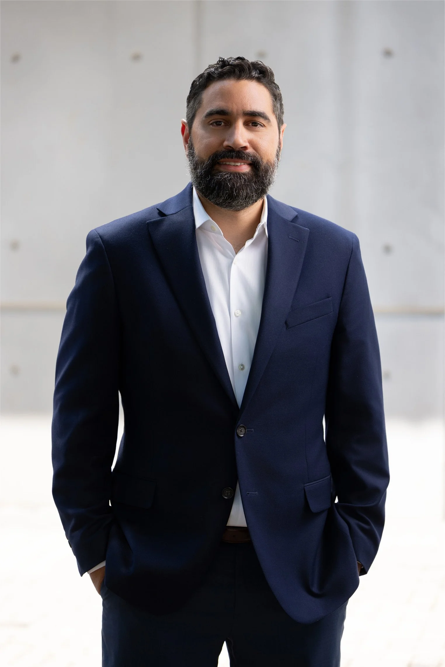 A man with dark hair and a beard wearing a navy blue suit and white shirt, standing with hands in pockets, in front of a light gray wall.