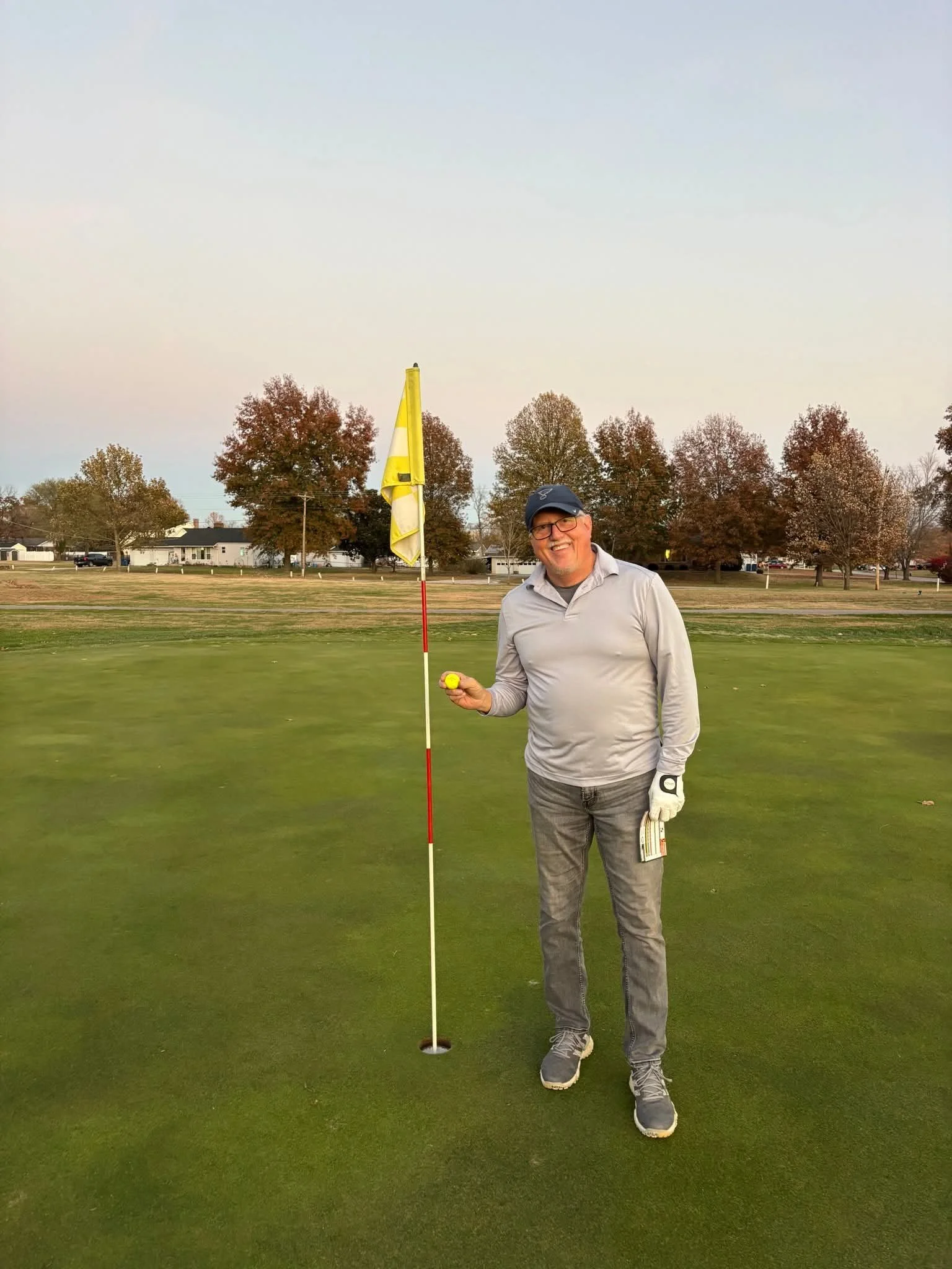 A man standing on a golf green holding a yellow golf ball, with a golf flag and hole nearby, in a park-like setting with trees and houses in the background.