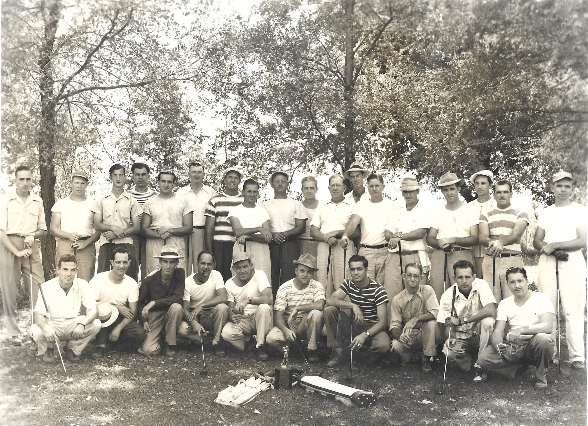 Group of men outdoors with trees in background, some holding golf clubs, some wearing hats, posing for a photo, with trophies and equipment on the ground.