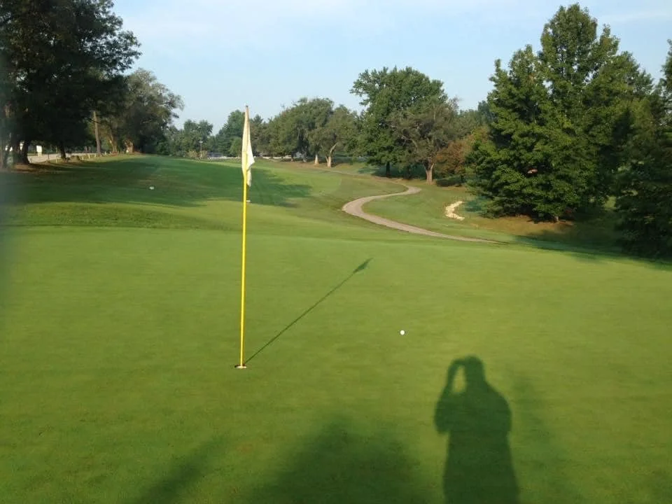 A golf course with a green, flagstick, and golf ball, surrounded by trees and a pathway, during daytime with clear skies.