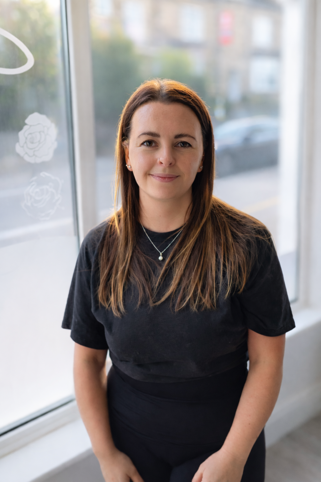 A woman with long brown hair, wearing a black shirt and layered necklaces, smiling at the camera inside a building near a window with flowers painted on it.