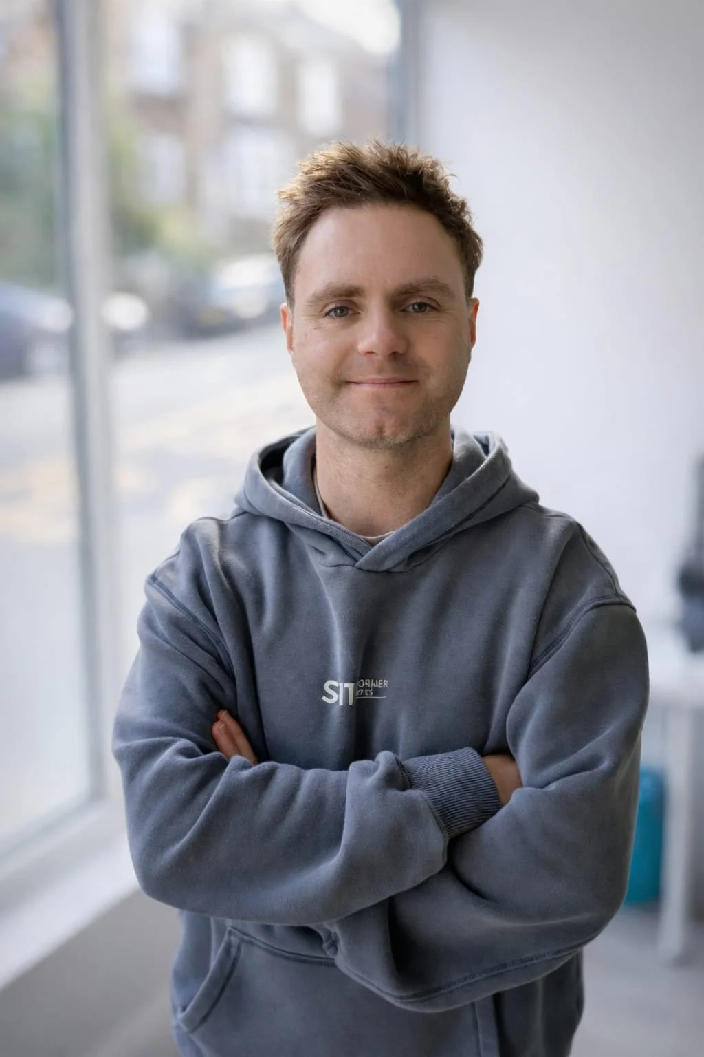 A young man with short, tousled brown hair and light skin, smiling slightly with eyes closed, stands indoors with arms crossed, wearing a gray hoodie with partially visible text on it.