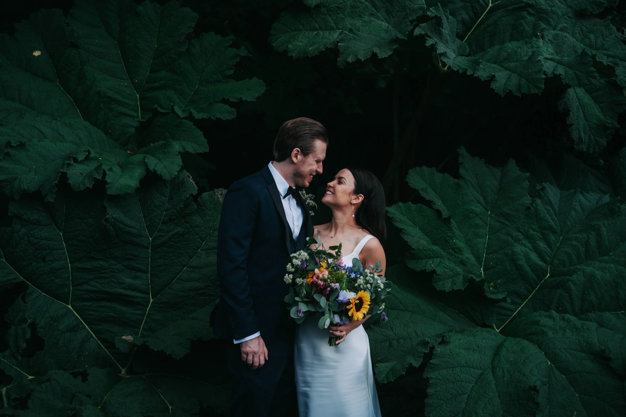 A bride and groom in wedding attire standing close together, smiling, with large green leaves in the background. The bride is holding a colorful bouquet of flowers.