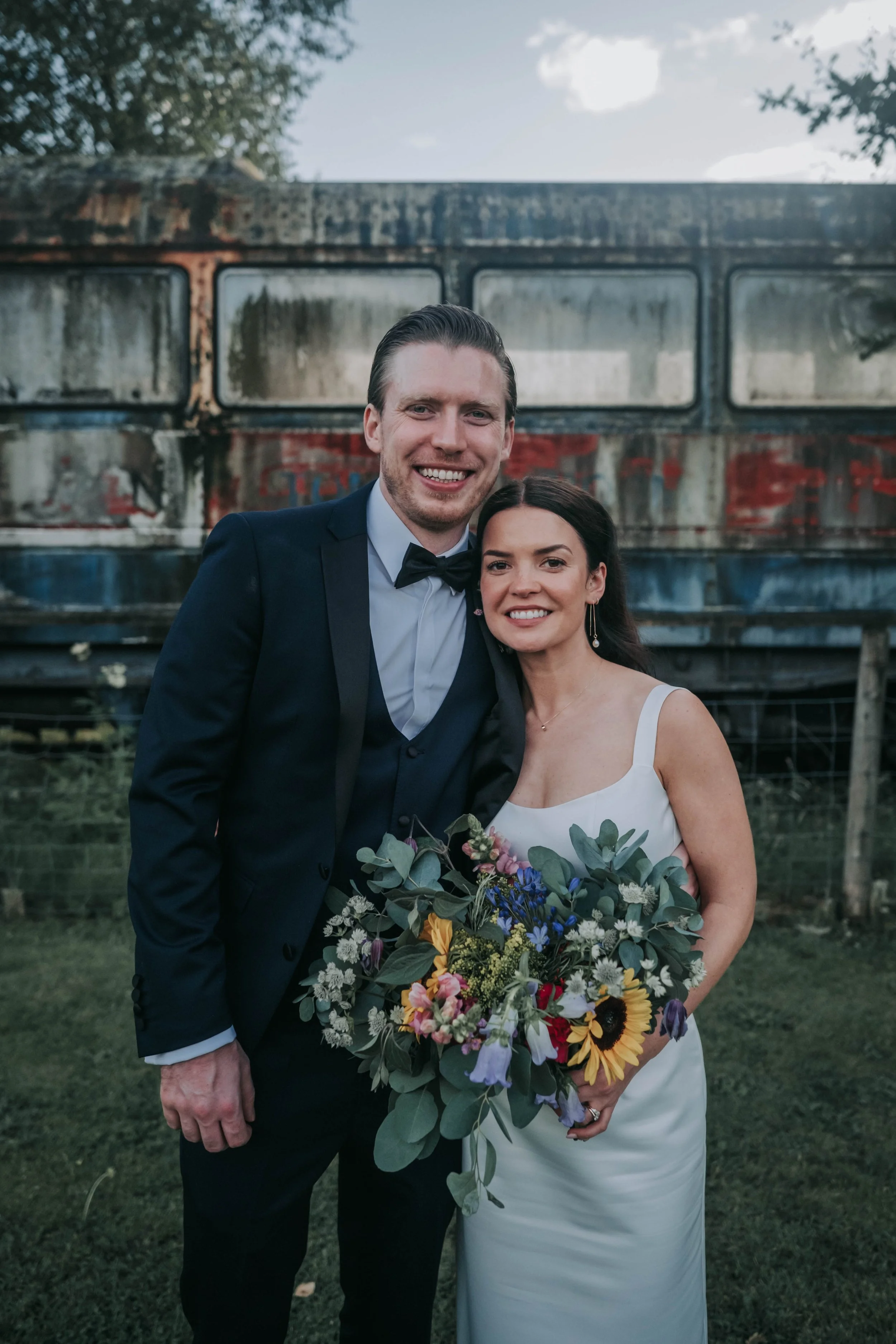 A happy couple on their wedding day posing outdoors in front of an old, rusted trailer. The groom is wearing a navy blue suit with a bow tie, and the bride is in a white wedding dress holding a colorful bouquet of flowers.