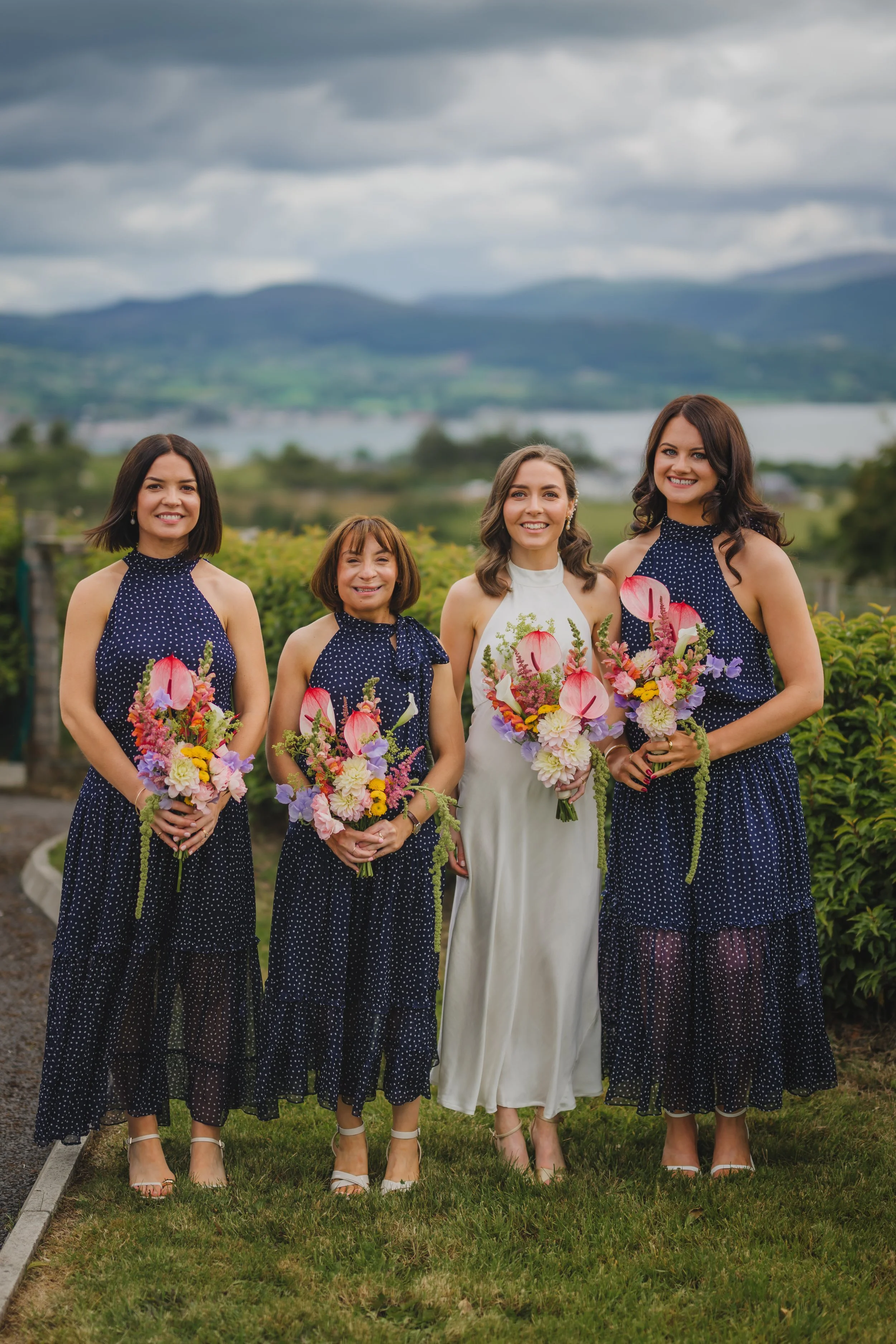 Four women standing outdoors holding colorful bouquets of flowers, with mountains and a lake in the background.