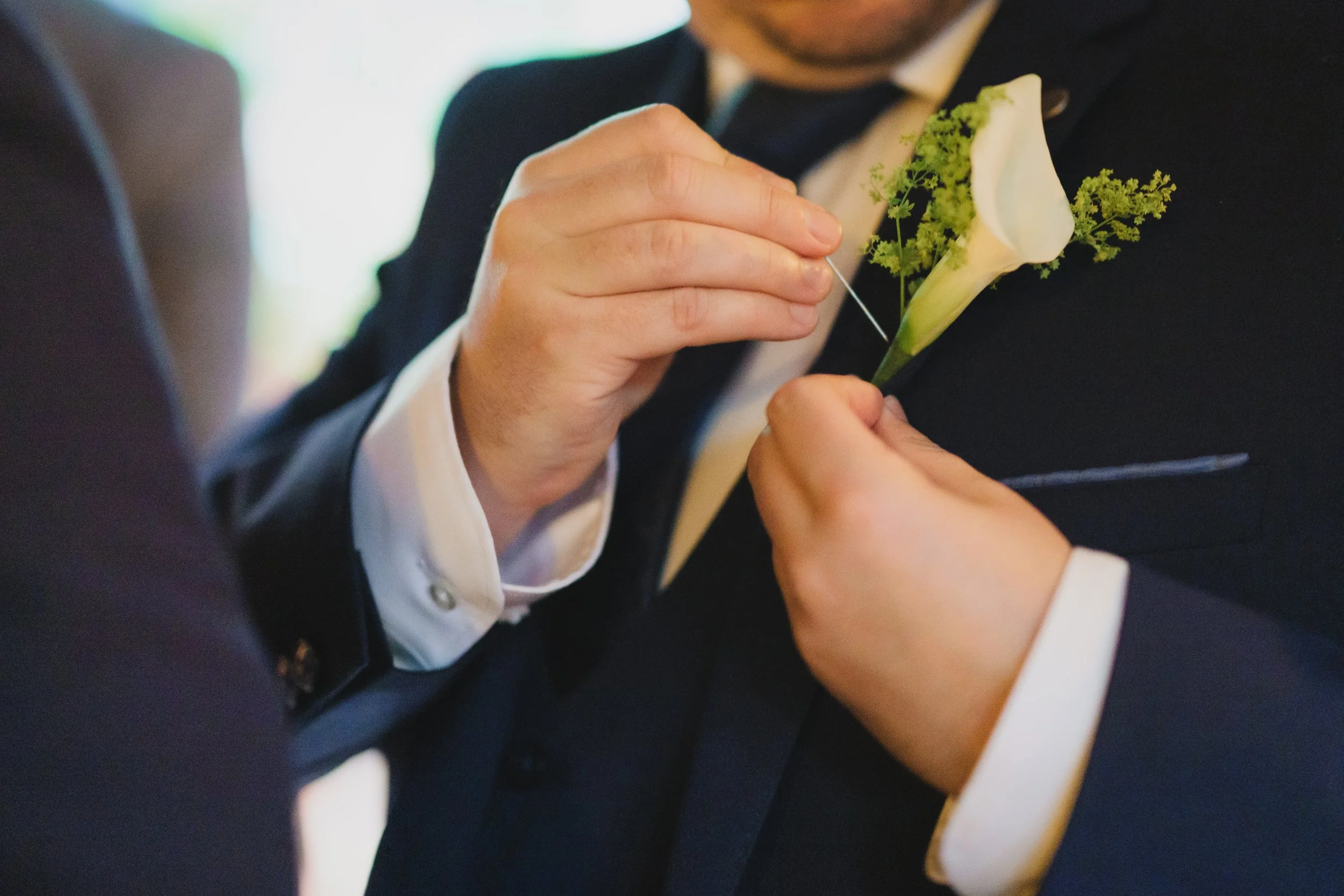 A person pinning a white calla lily and green foliage buttonhole onto a man's suit jacket at a formal event.