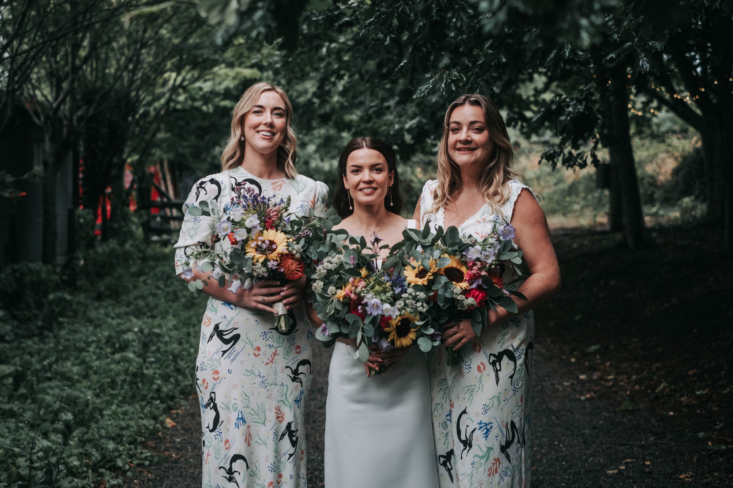 Three women in a wooded outdoor setting, dressed in white and holding large colorful bouquets, smiling at the camera.
