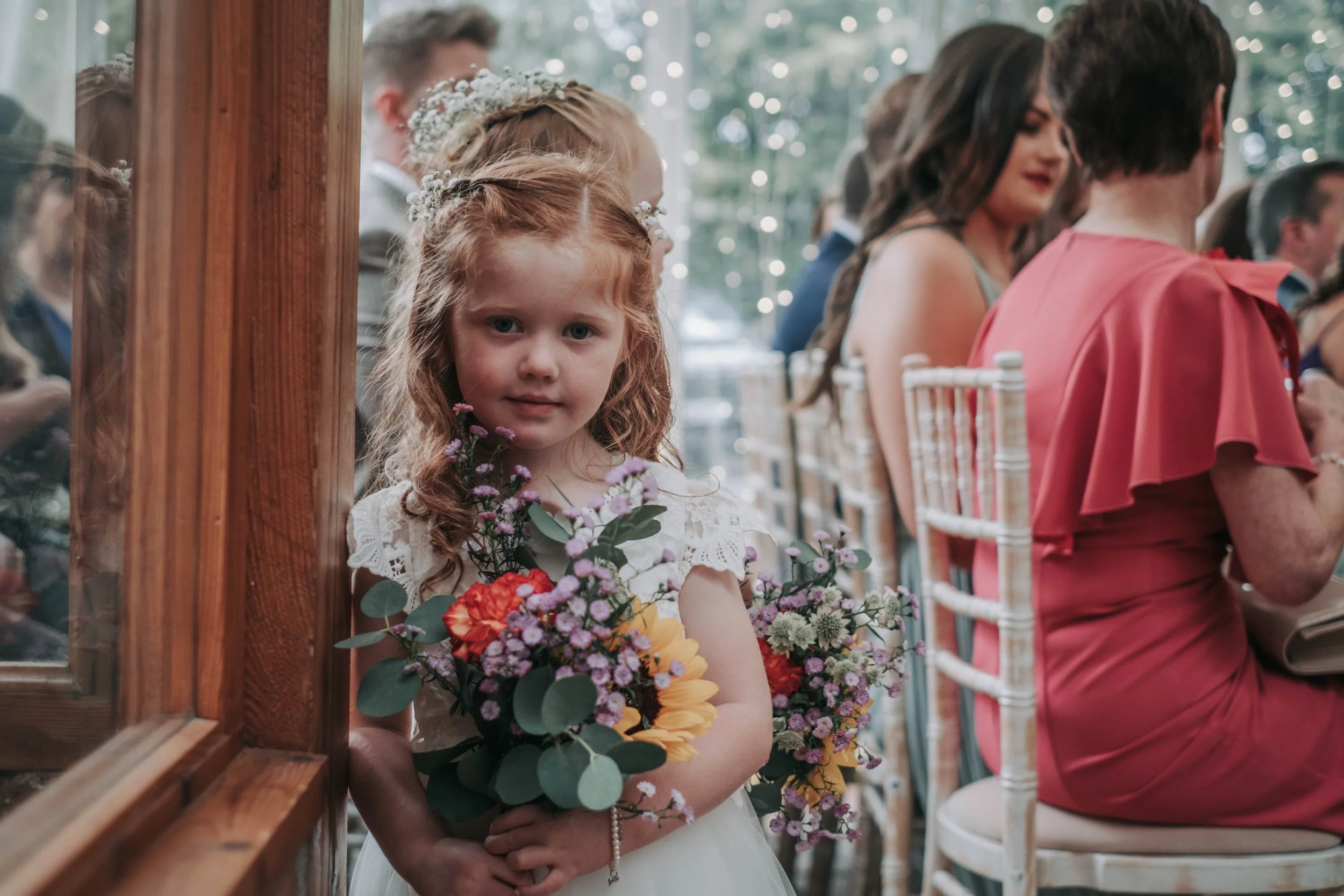 A young girl with red hair holding a bouquet of colorful flowers at a wedding or formal event, sitting among seated guests.
