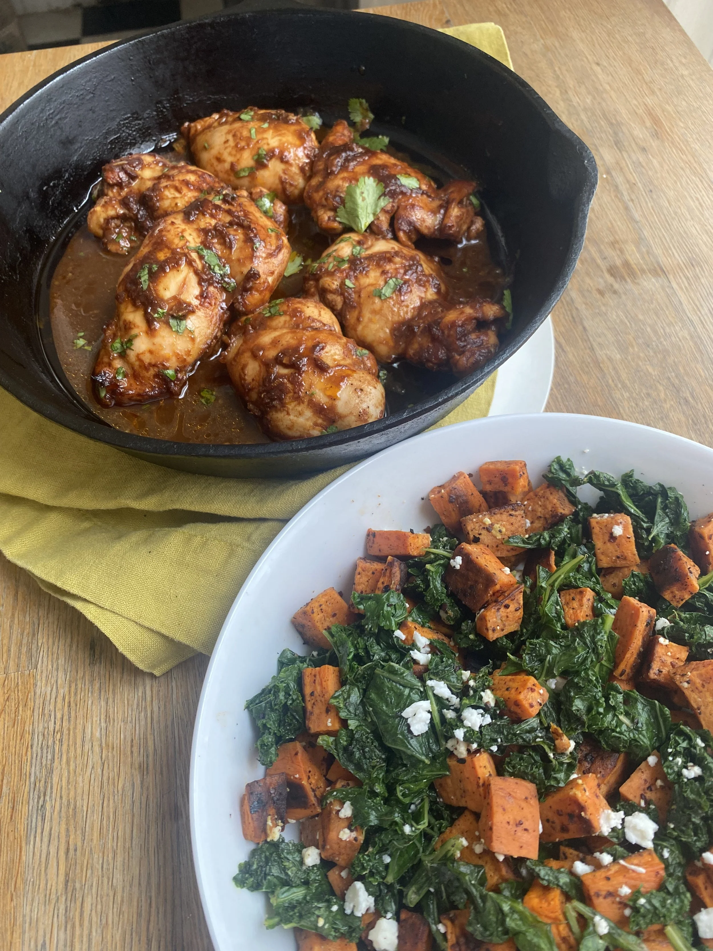 A close-up of a dish of cooked chicken drumsticks covered in barbecue sauce, garnished with cilantro, in a black cast iron skillet. In the foreground, there is a white plate with roasted sweet potatoes and kale, topped with crumbled cheese.