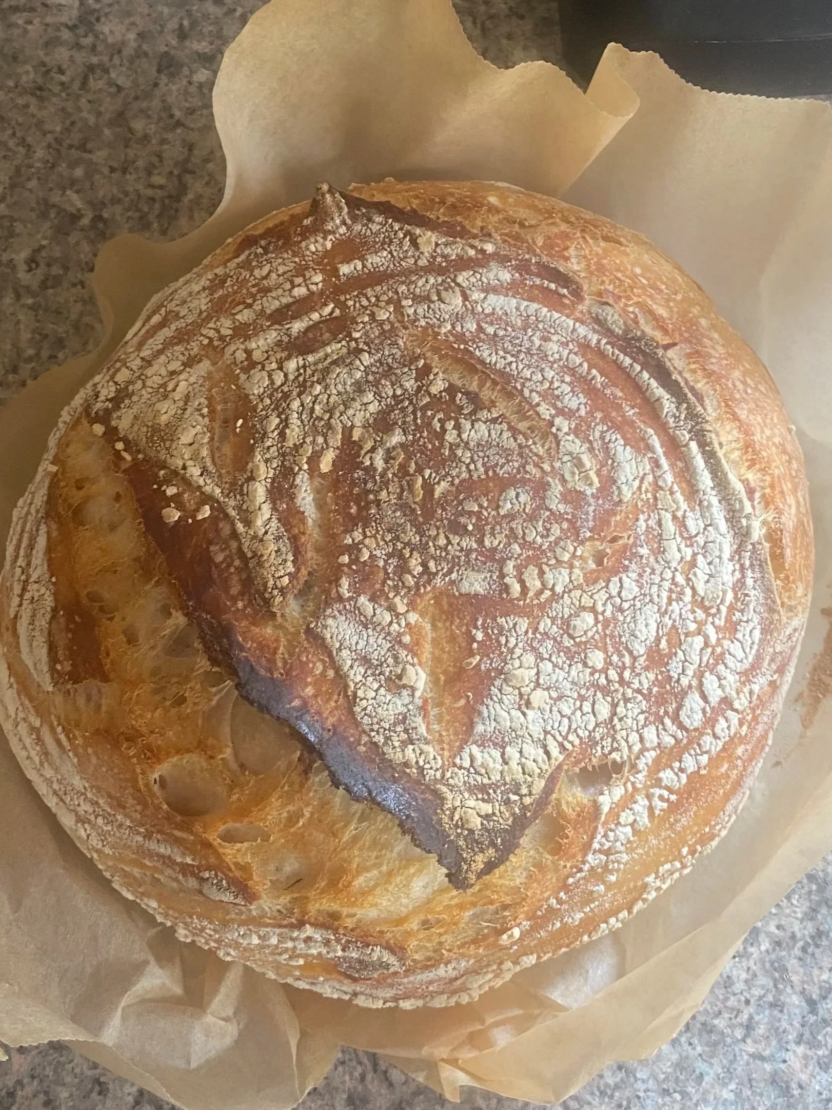 A round loaf of rustic bread with a cracked crust, dusted with flour, resting on parchment paper.