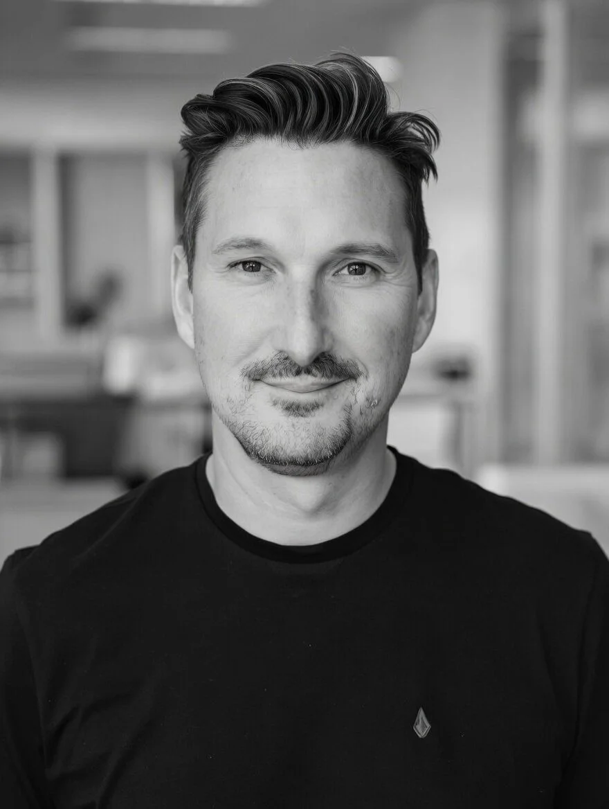 A black and white portrait of a man with styled hair, a slight smile, and facial hair, wearing a dark t-shirt with a small logo on the chest. The background is blurred.