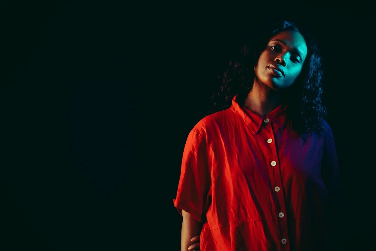 Young woman with curly hair wearing a red button-up shirt against a dark background with blue and green lighting.