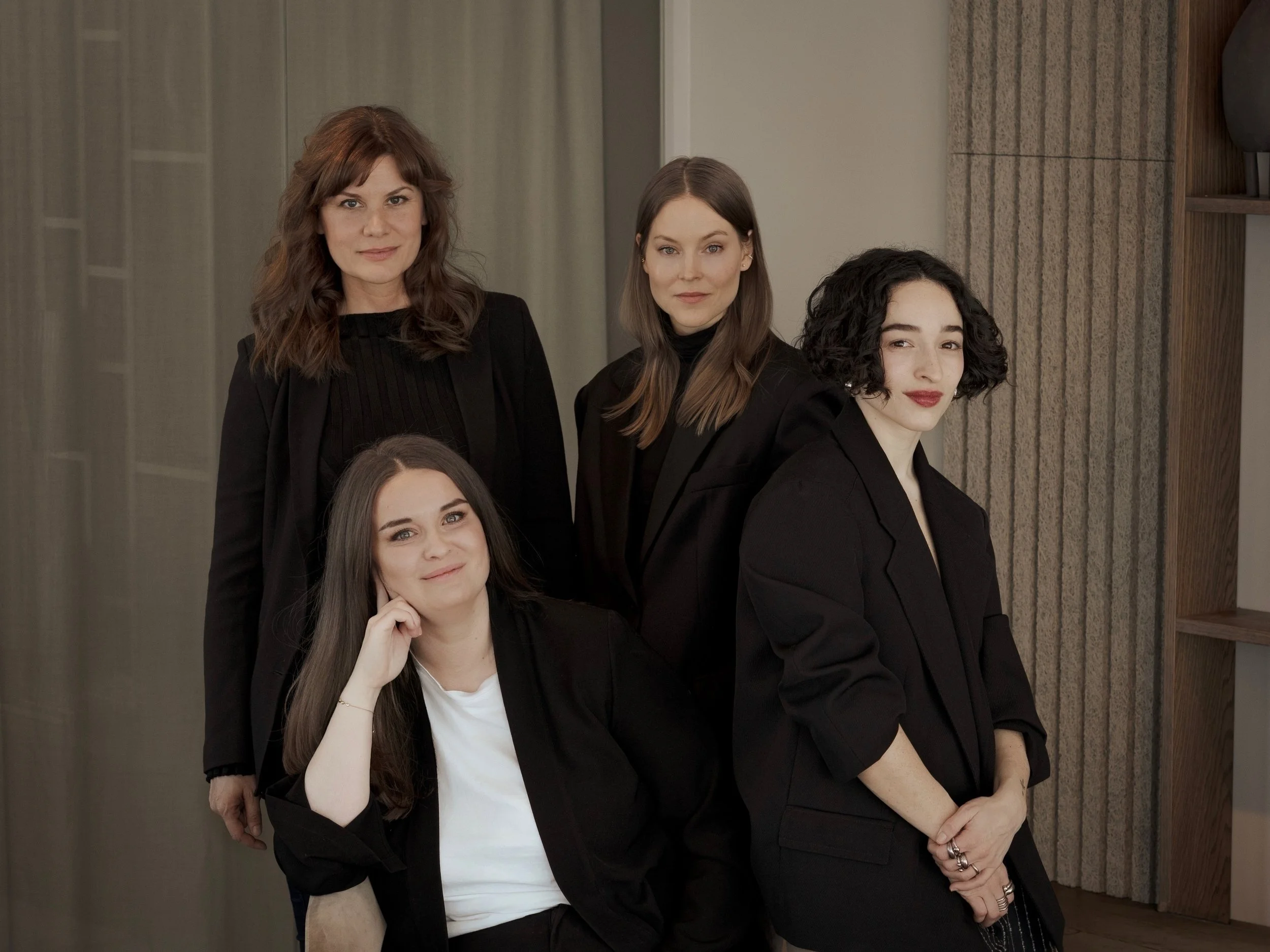 Five women, dressed in black, pose together indoors against a neutral background with a curtain and wooden shelving. They are arranged with two women standing in the back and three in front, one sitting and the others standing. They look at the camera with confident expressions.