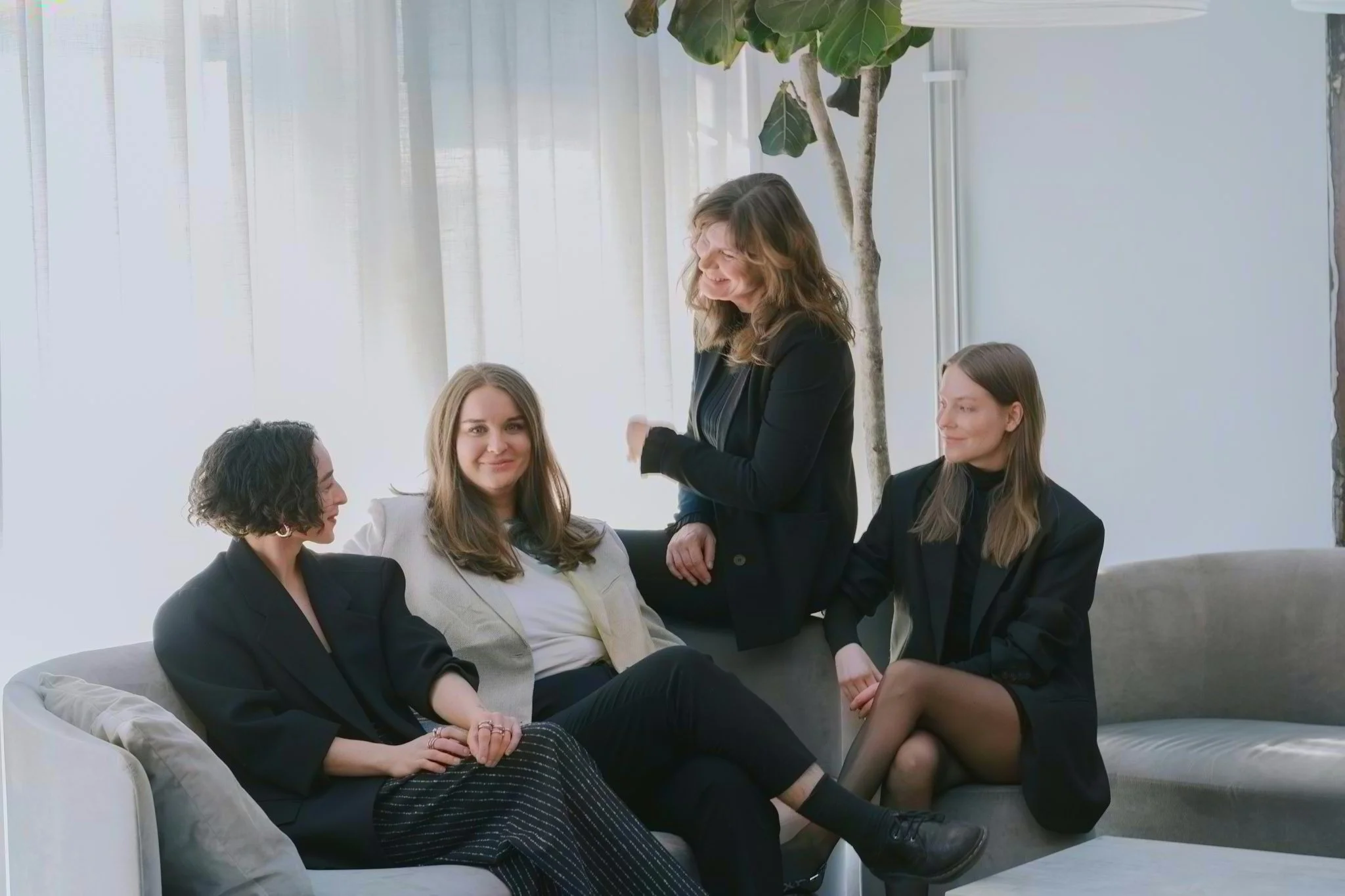 Four women engaging in a conversation in a bright room with large windows and a tall plant in the background. Three are seated on a sofa, and one is standing, smiling.
