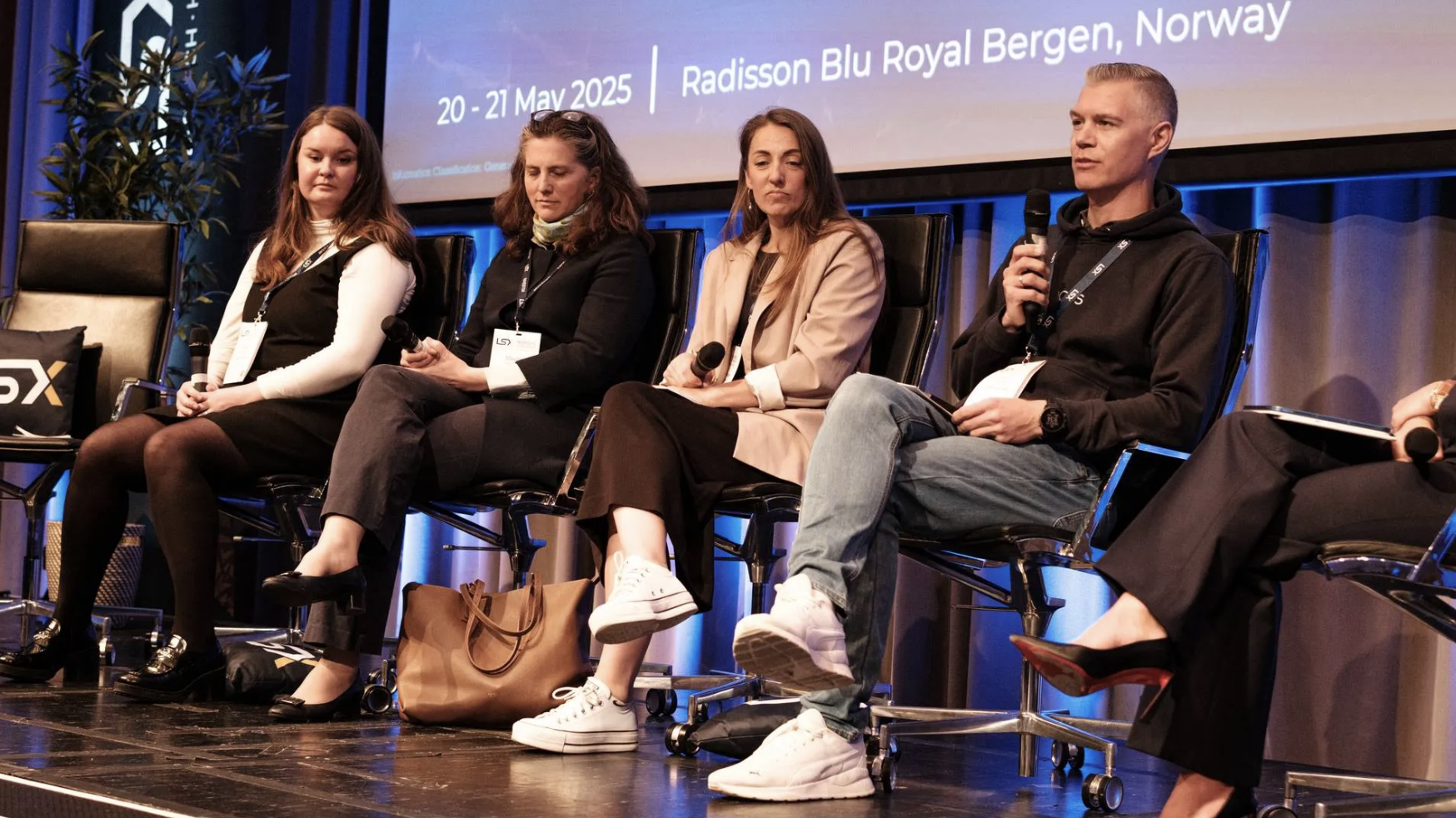 Five people sitting on a stage during a panel discussion at a conference, with a large screen behind them displaying conference details and dates.