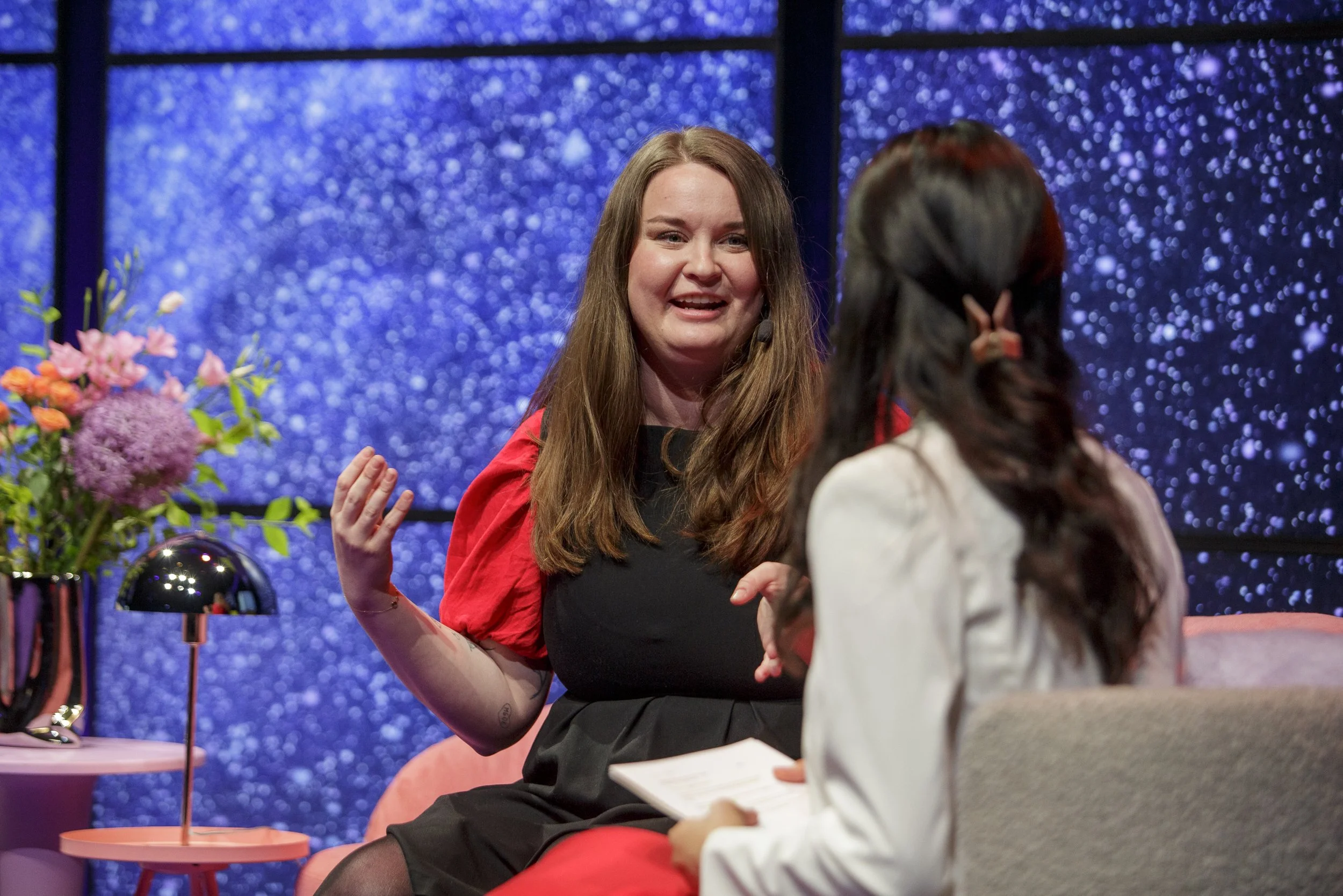 Two women having a conversation on a stage or set with a blue starry background. One woman has long brown hair and is wearing a black and red outfit, and the other woman has dark hair with an orange hairpin, holding a piece of paper.