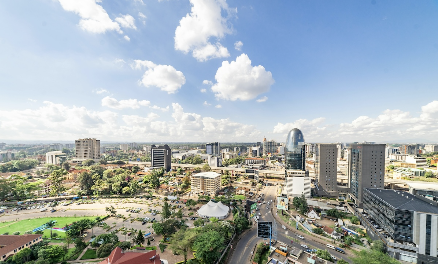 A cityscape with tall modern buildings, a green park in the foreground, and a partly cloudy blue sky overhead.