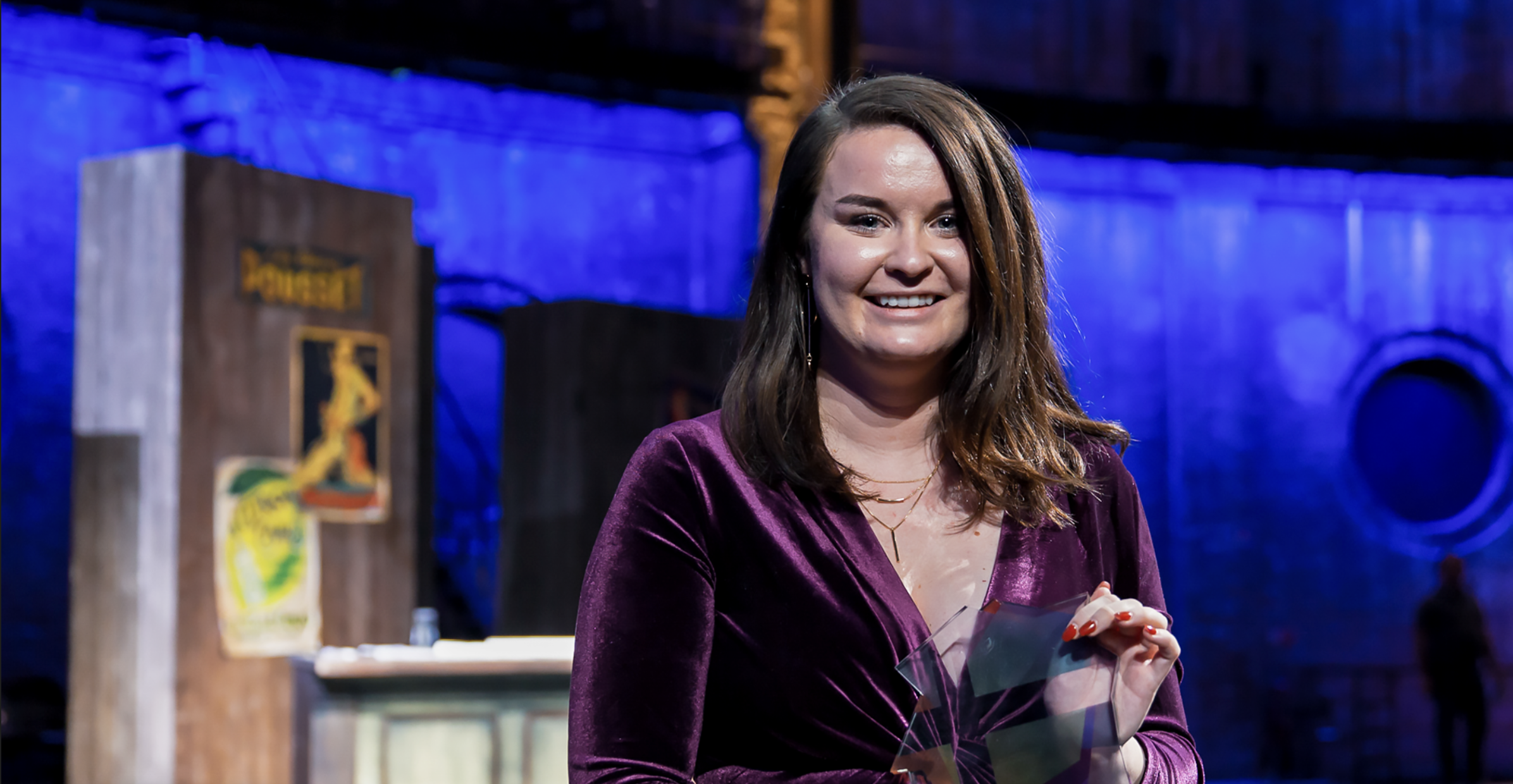 A woman with shoulder-length brown hair, wearing a purple velvet dress, smiling and holding a glass award in front of a dark, industrial-style backdrop with blue lighting.