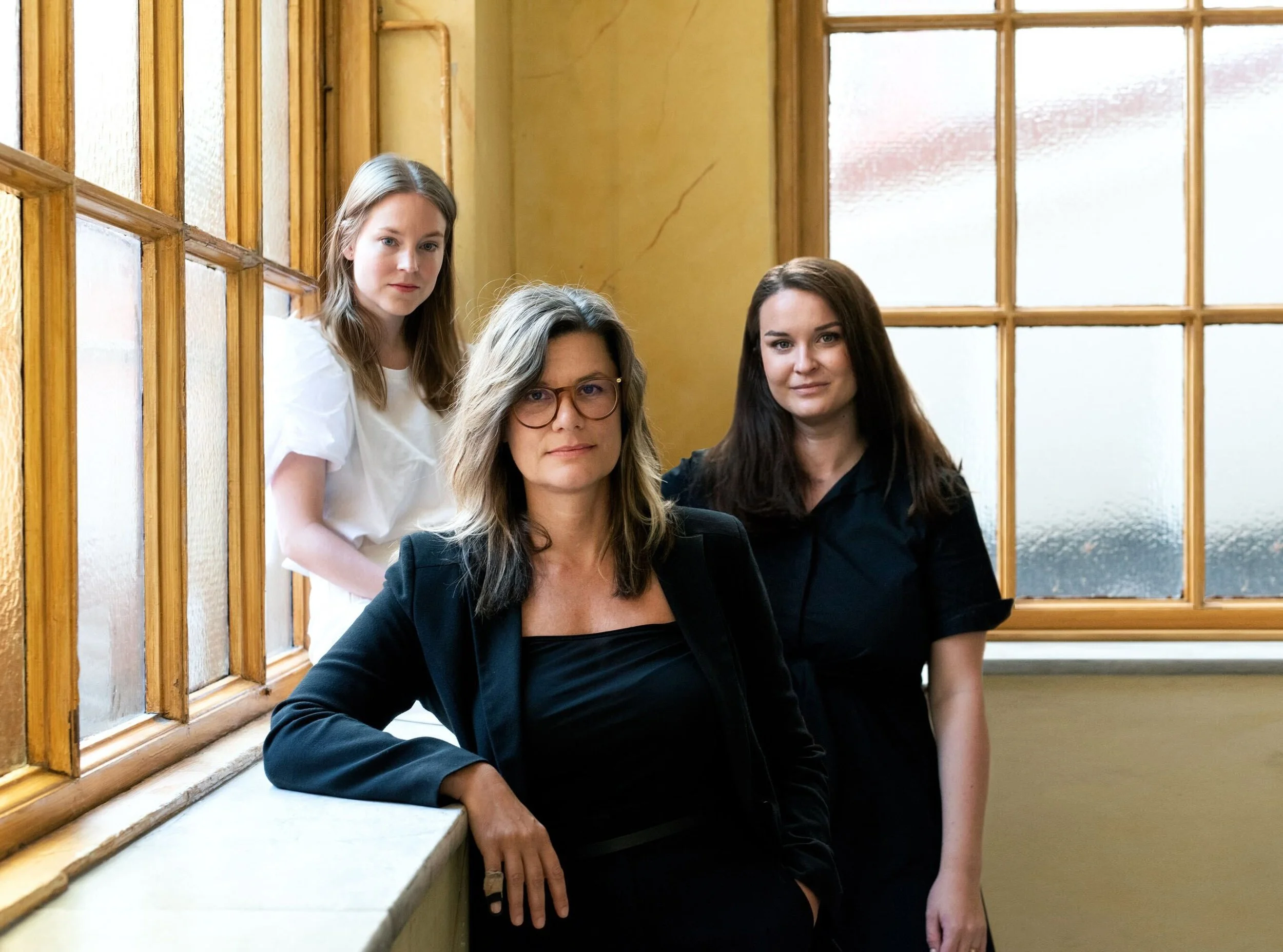 Three women standing indoors in front of frosted glass windows, with the woman in the front wearing glasses and a black jacket, and the women in the back wearing black and white blouses.