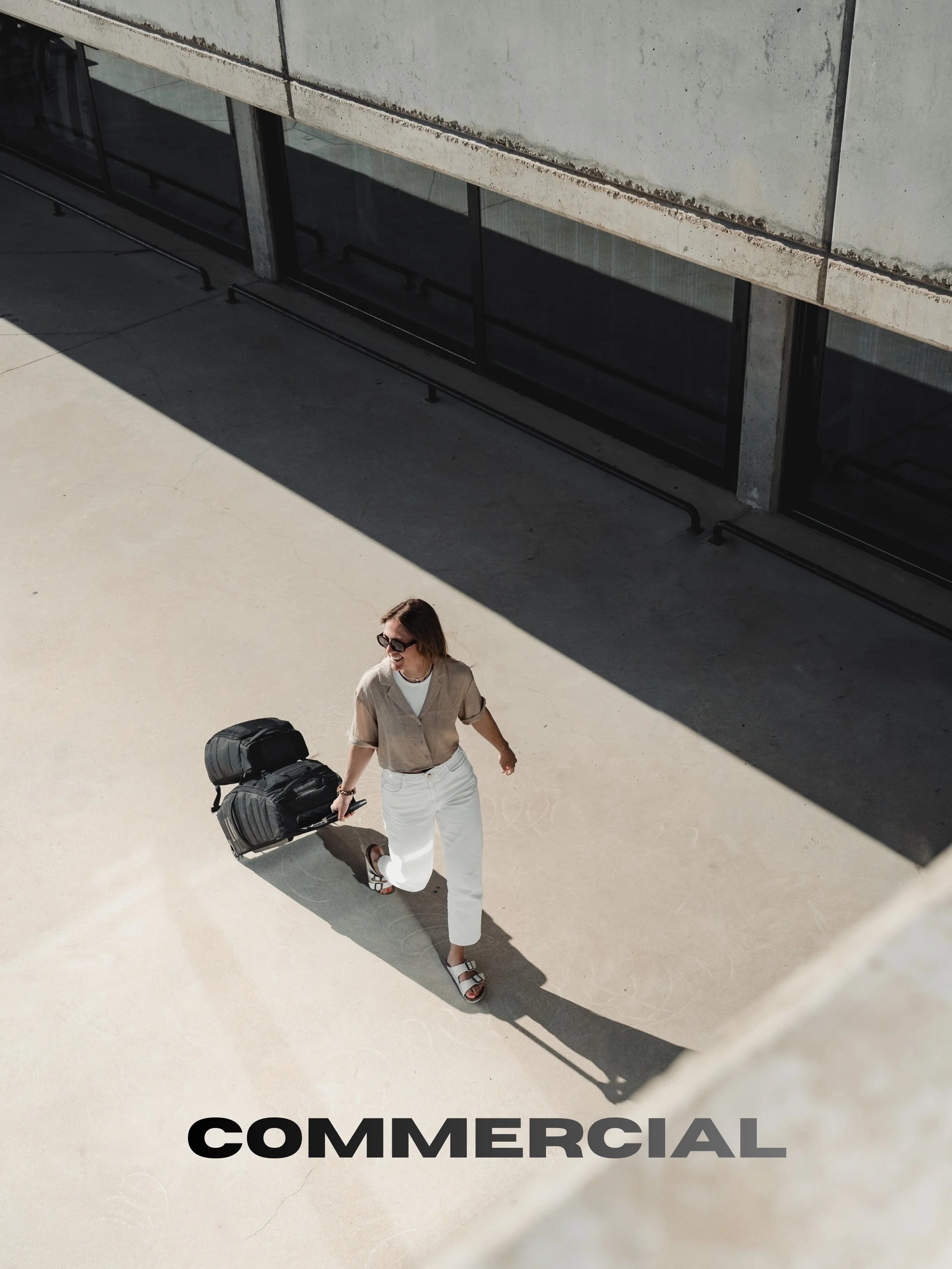 A woman with sunglasses, wearing a beige shirt and white pants, walking with a rolling suitcase in an outdoor urban setting with modern building and shadow.
