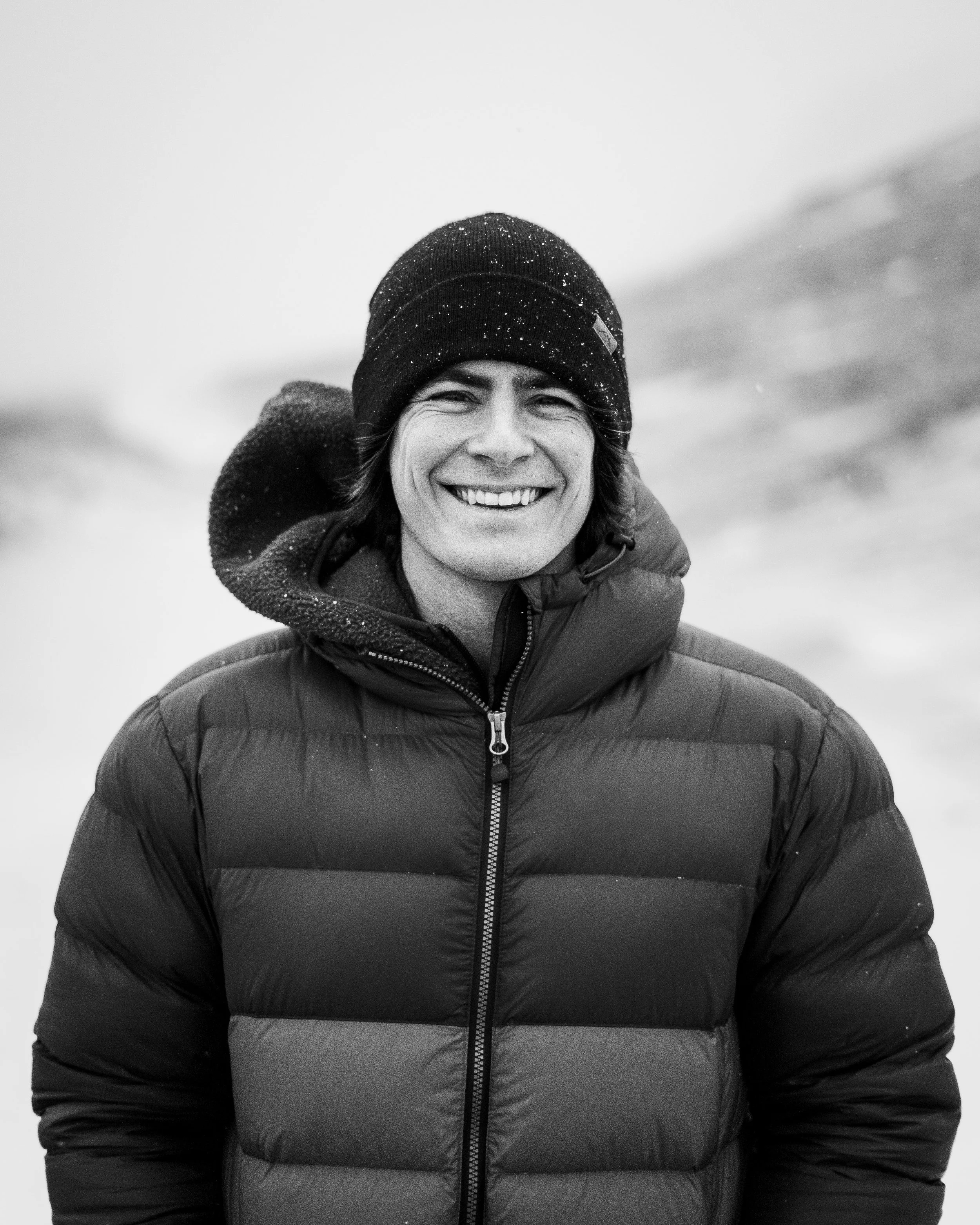 A smiling man in a winter jacket and knit hat outdoors on a snowy day.