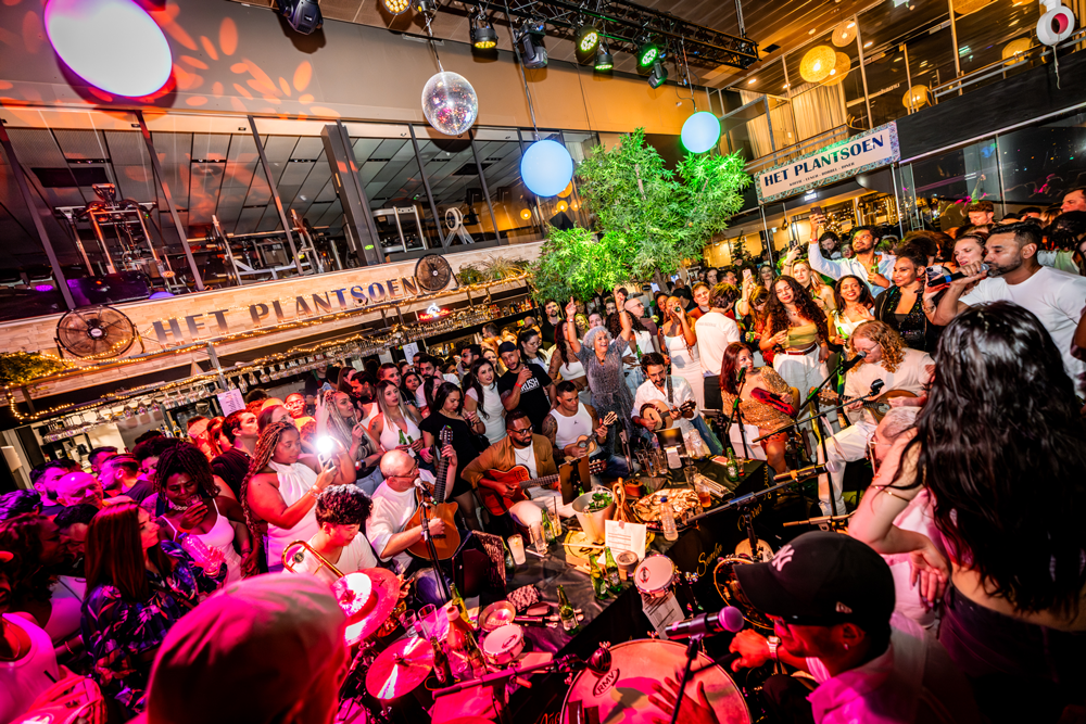 A lively indoor party or concert with a large crowd of people, some playing guitars and drums, in a decorated venue with colorful lighting, green plants, and disco balls.
