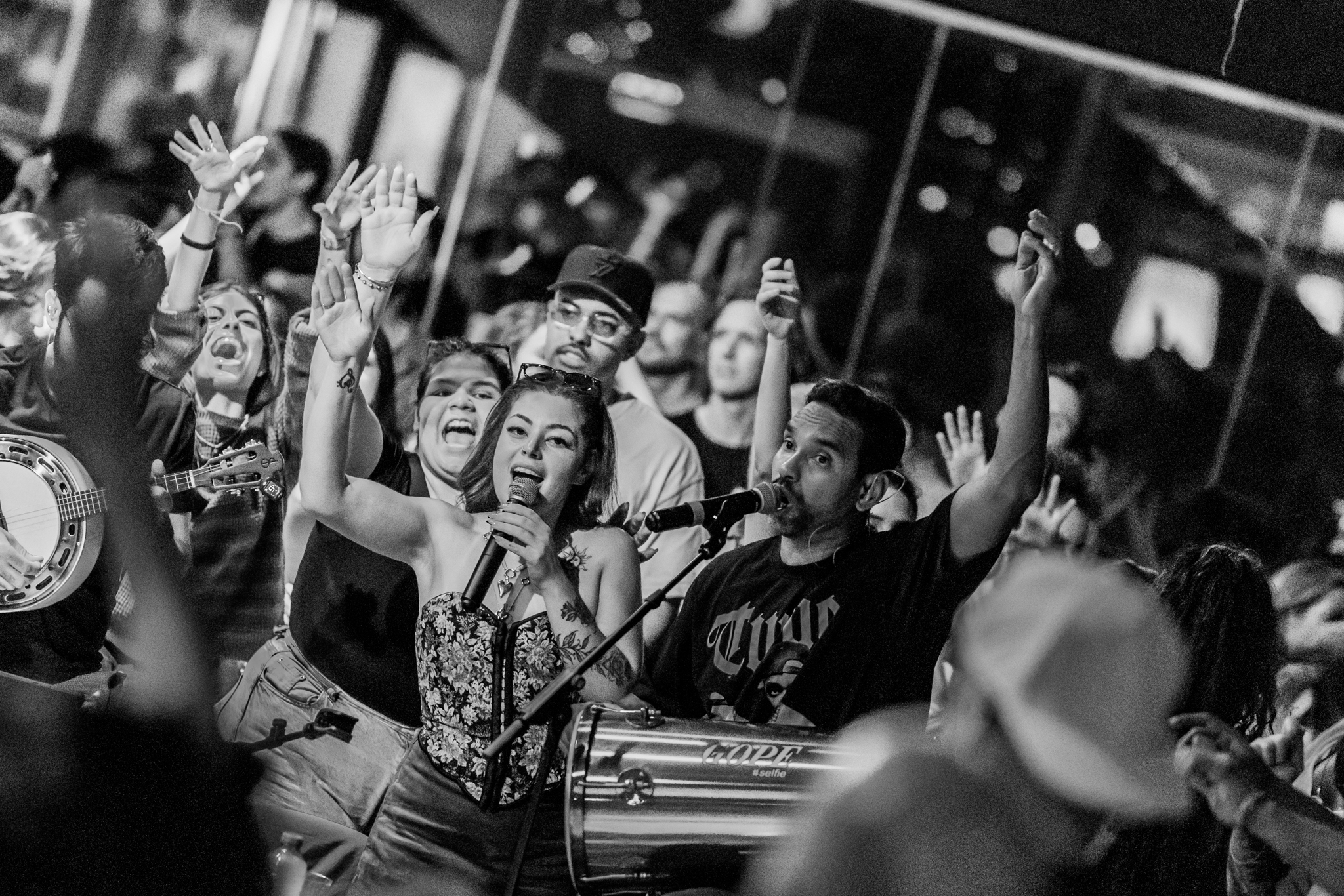 A group of people at a lively gathering, some holding musical instruments, with two central figures singing into microphones, raising their hands, surrounded by cheering crowd.