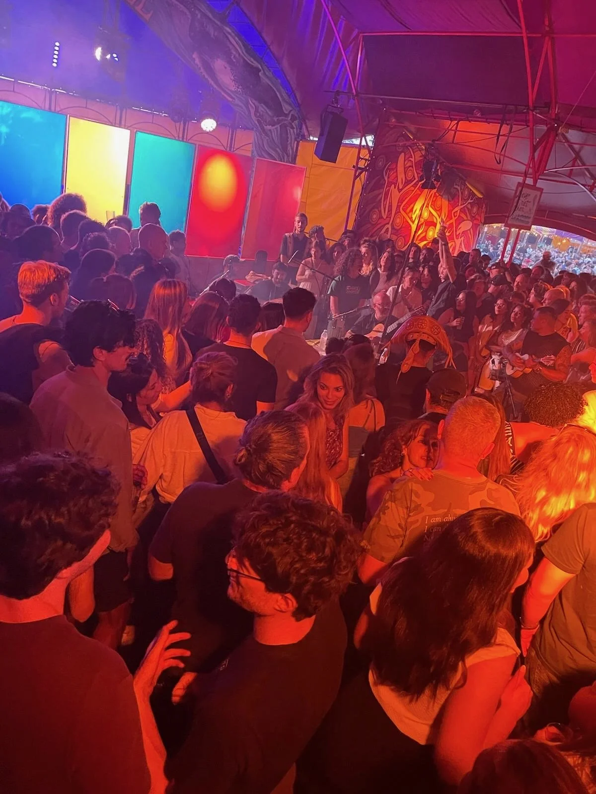 Crowd of people under a tent with colorful abstract artwork and lighting listening and vibing with Samba Roots the band that brings Brazilian Culture to Europe
