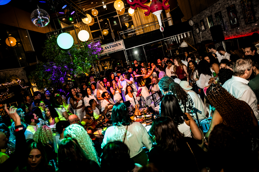 Crowd of people dancing and enjoying live Roda De Samba from SambaRoots in a lively indoor party with colorful lights and decorations
