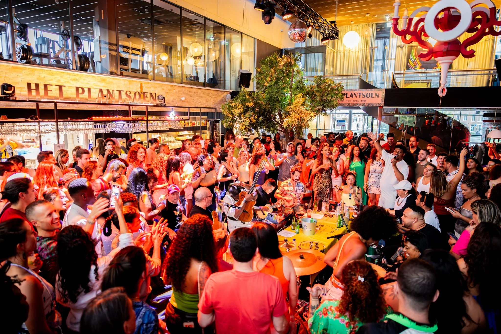 A lively indoor concert with a large crowd, musicians playing guitars, a singer with a microphone, and people dancing and enjoying the music under colorful lighting and decorations.
