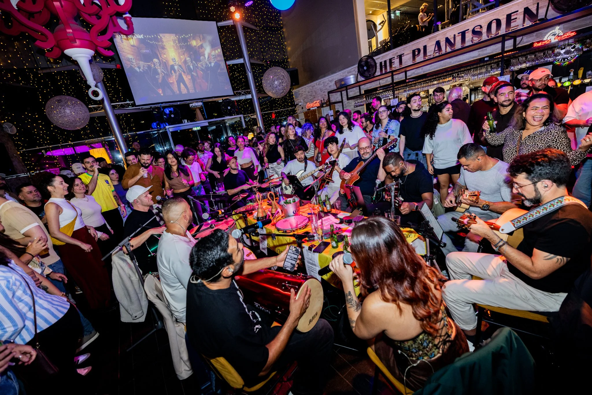 Crowd of people gathered around a live band performing at an indoor venue, with colorful lighting and a large screen displaying animated characters in the background.