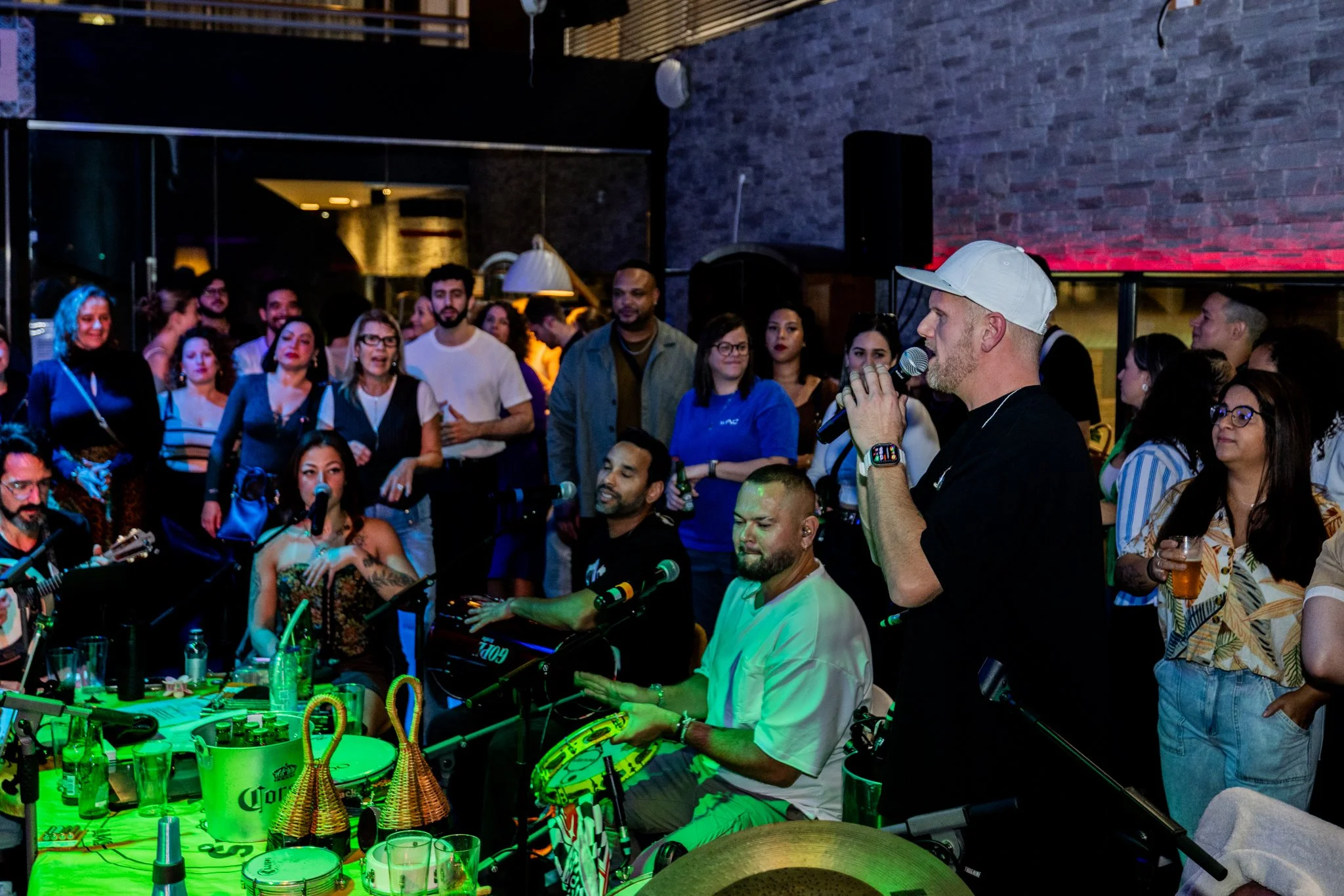 A man in a white cap and black shirt is speaking into a microphone at a lively indoor gathering with musical performers and an audience.