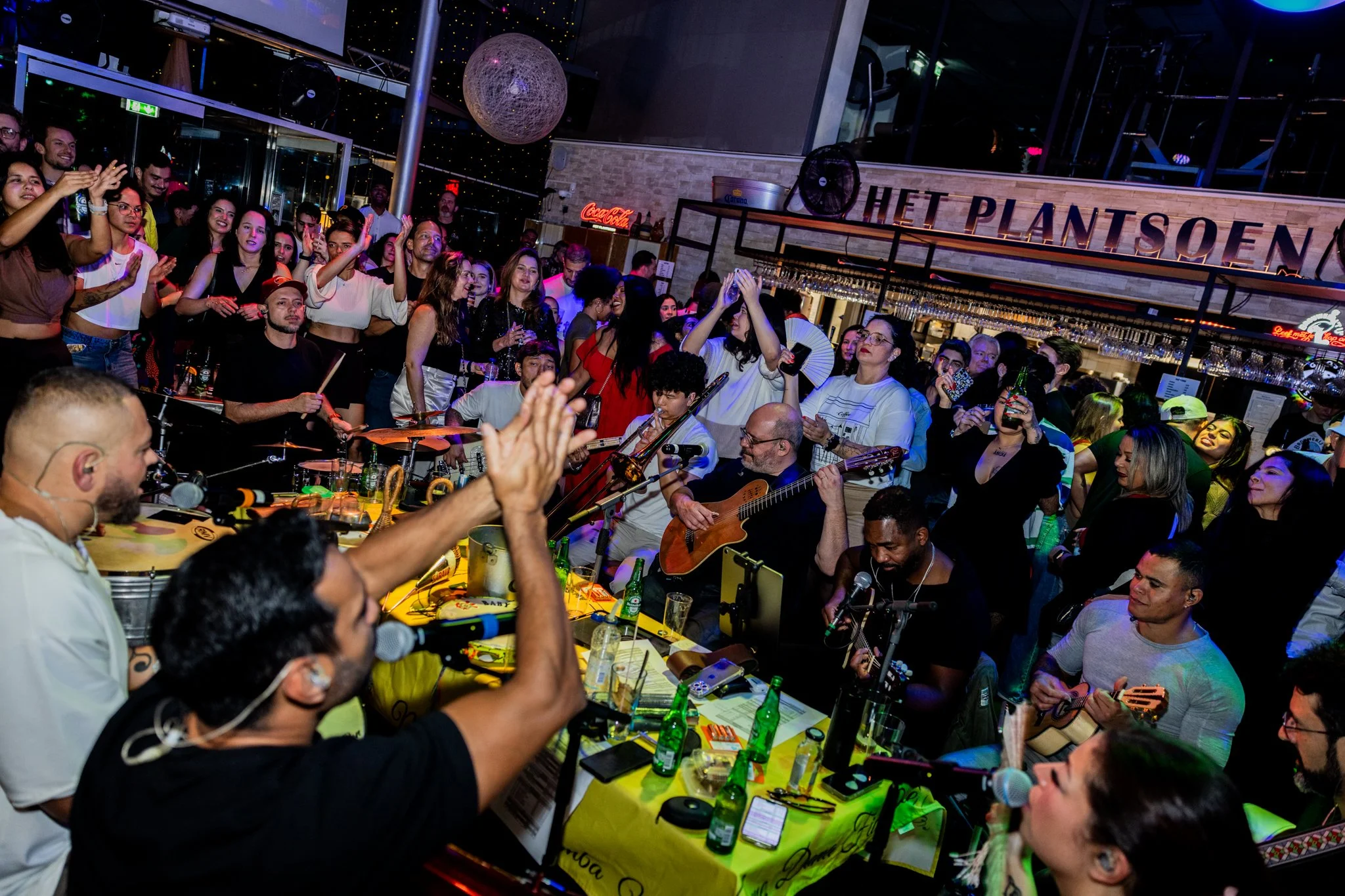 A lively indoor concert with musicians playing guitar and percussion, surrounded by a crowd of enthusiastic audience members, some clapping and others taking photos, with colorful lighting and a sign reading 'Het Planten' in the background.