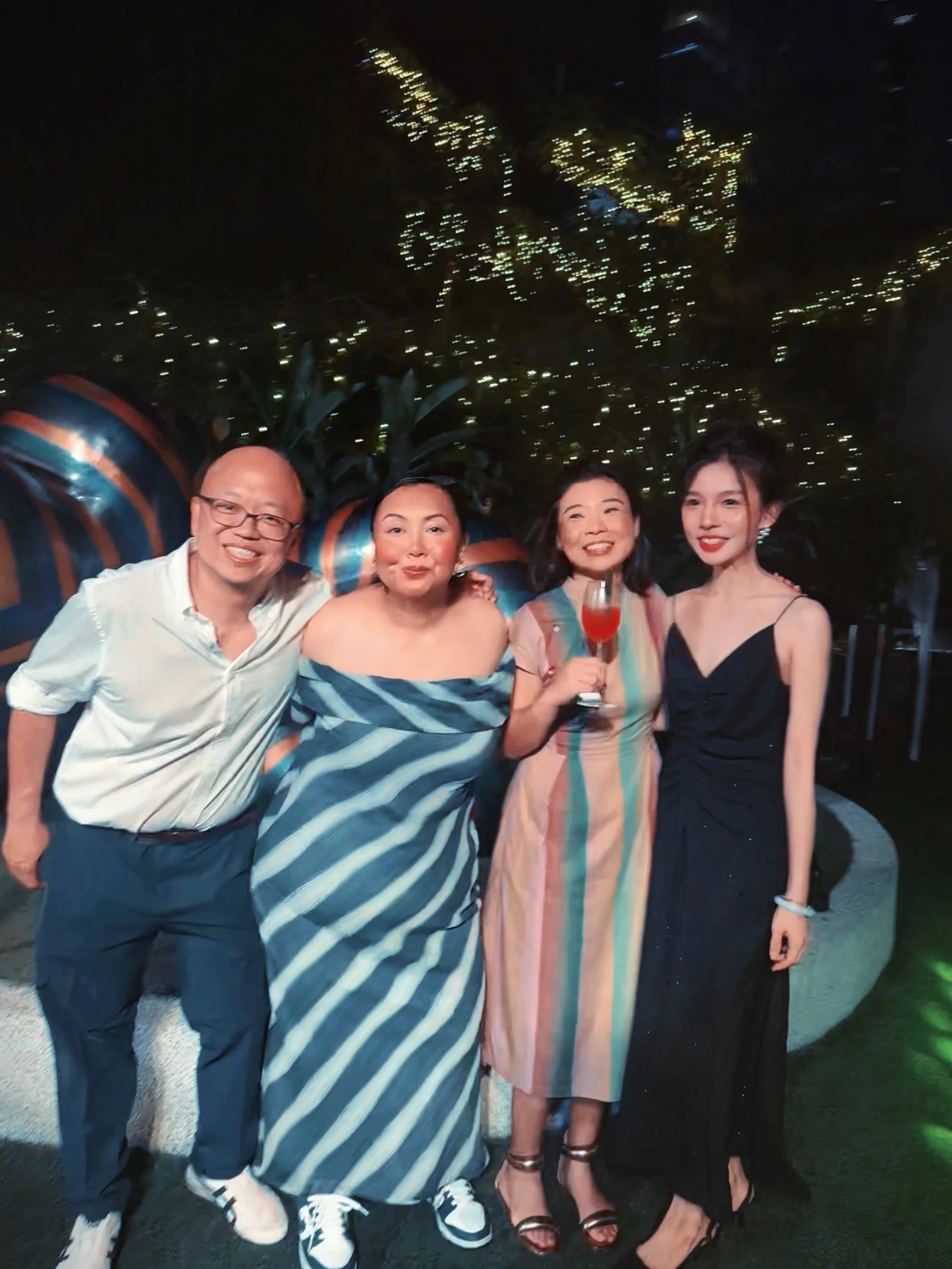 Group of four people at nighttime outdoor event, three women and one man, smiling and posing in front of a decorative background with lights and large wrapped objects, some holding drinks, dressed in stylish outfits.