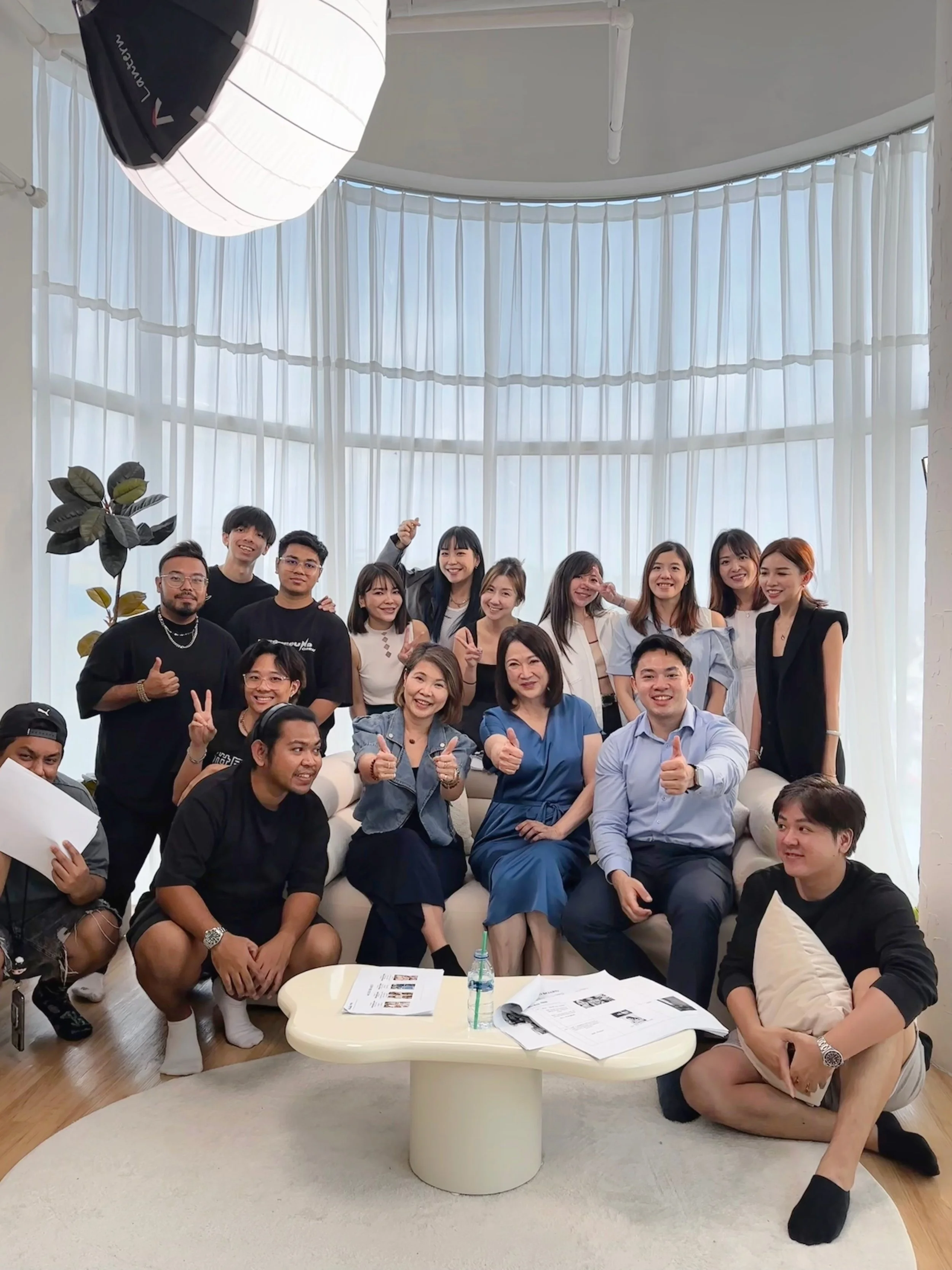 Group of people gathered in a well-lit room with large windows and white curtains, smiling and making various gestures, seated and standing around a white coffee table with papers and a bottle on it.