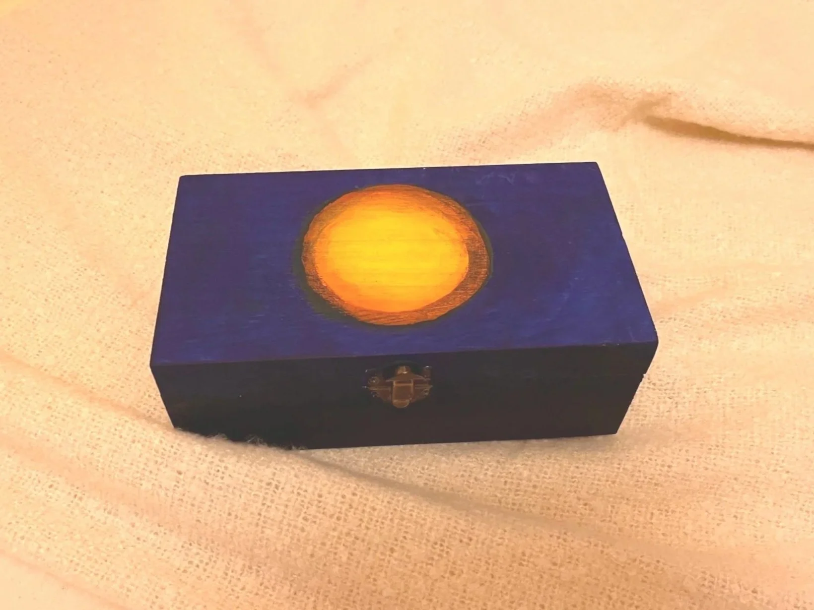 A small, dark blue box with a latch, featuring a glowing yellow-orange circle resembling a sun or moon on the top surface, placed on a light-colored fabric surface.