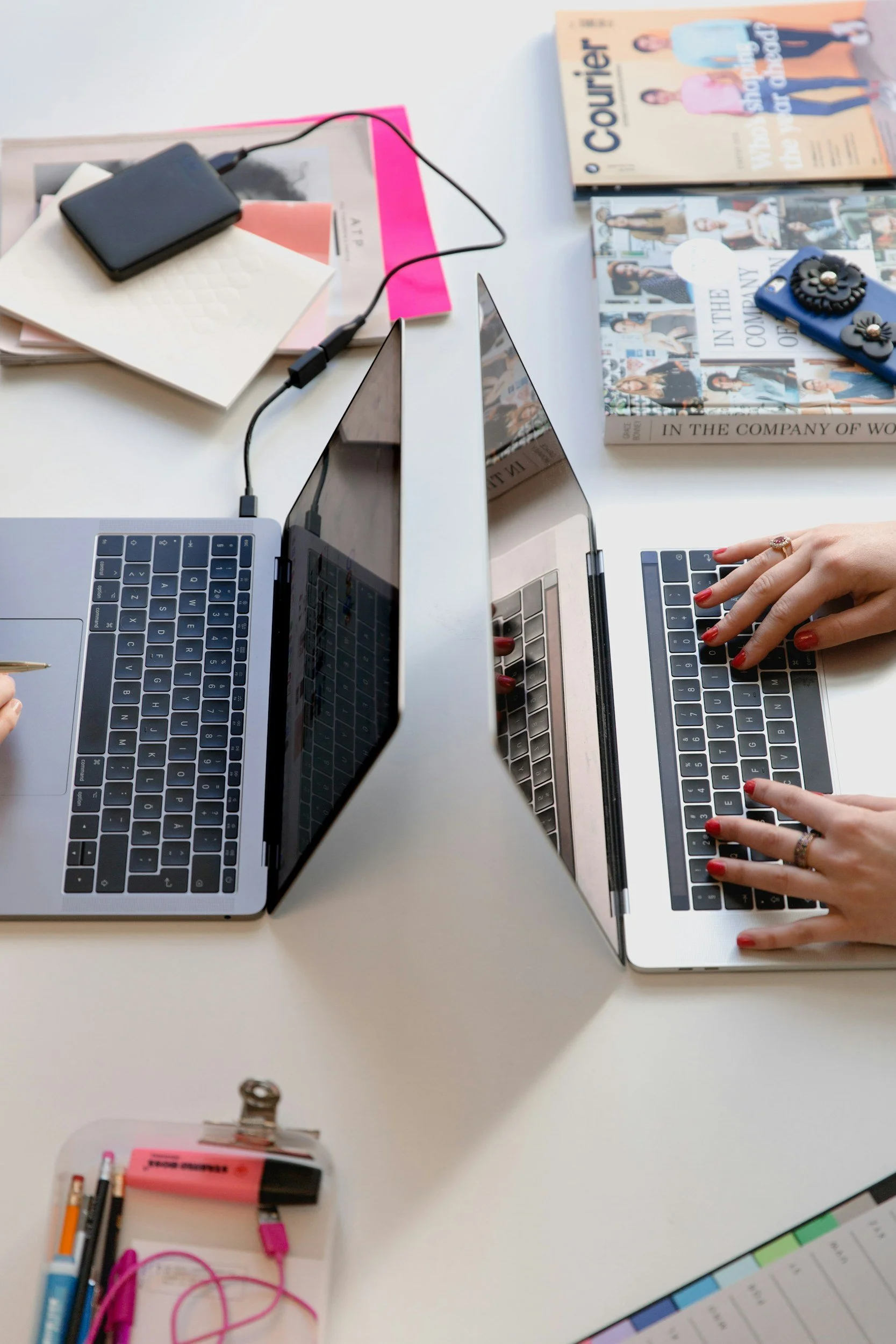 Two laptops on a white desk with two people typing on each keyboard, various notebooks, magazines, and office supplies scattered around.