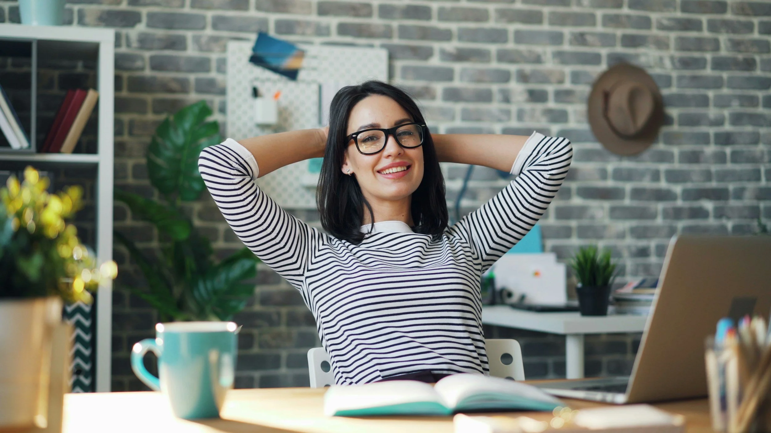 A woman with glasses sitting at a desk with her hands behind her head, smiling in a modern office with a gray brick wall, shelves with books, a hat on the wall, and plants.