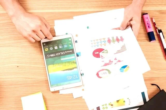 Person reviewing printed documents with colorful charts and graphs on a wooden table, with a smartphone, highlighters, and other office supplies nearby.