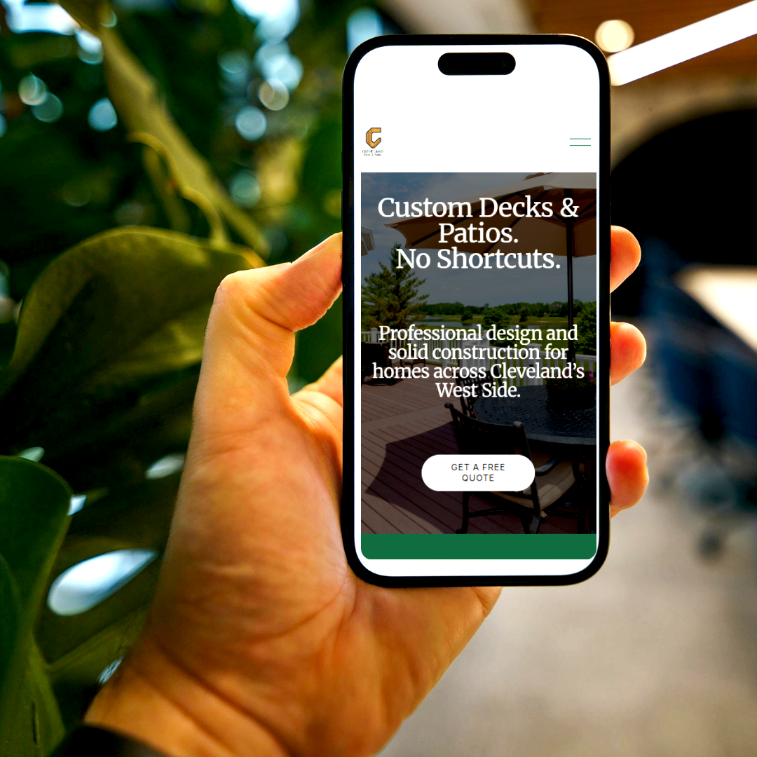 Hand holding a smartphone displaying a website for custom decks and patios, with a background of outdoor furniture and trees.