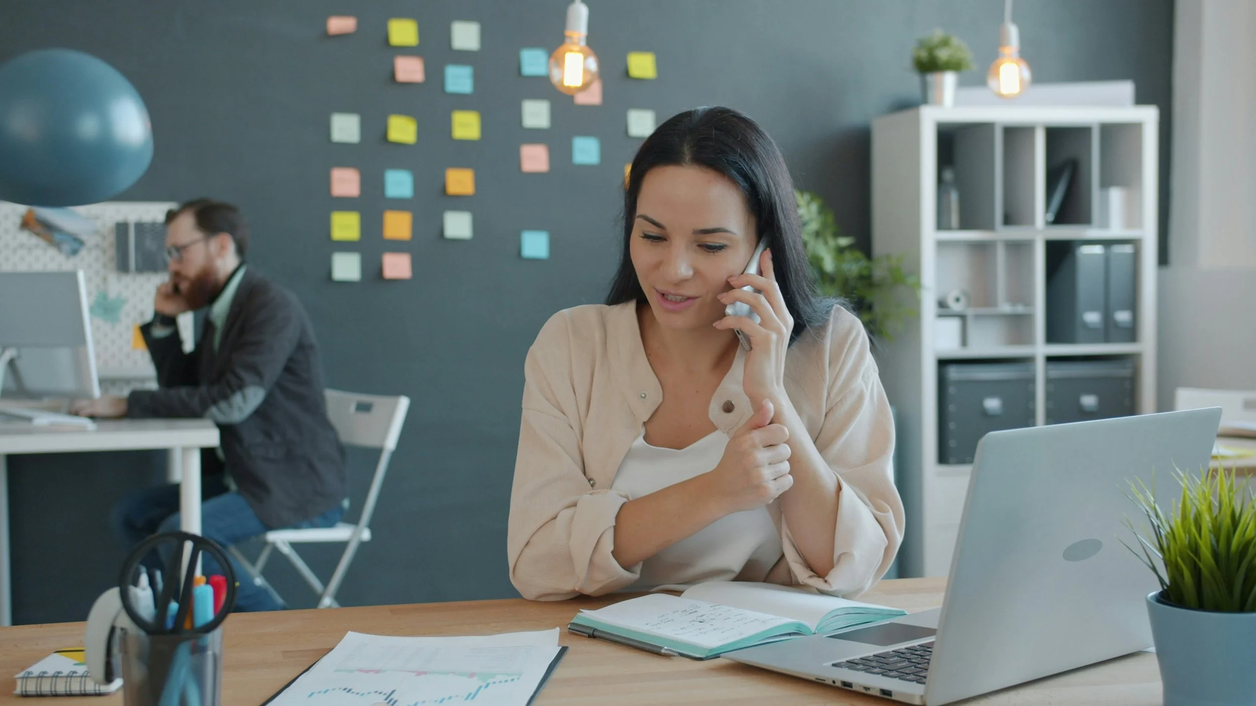 A woman talking on the phone at her desk with a laptop, notebook, and documents, in a modern office with colorful sticky notes on a wall and a man working at a computer in the background.