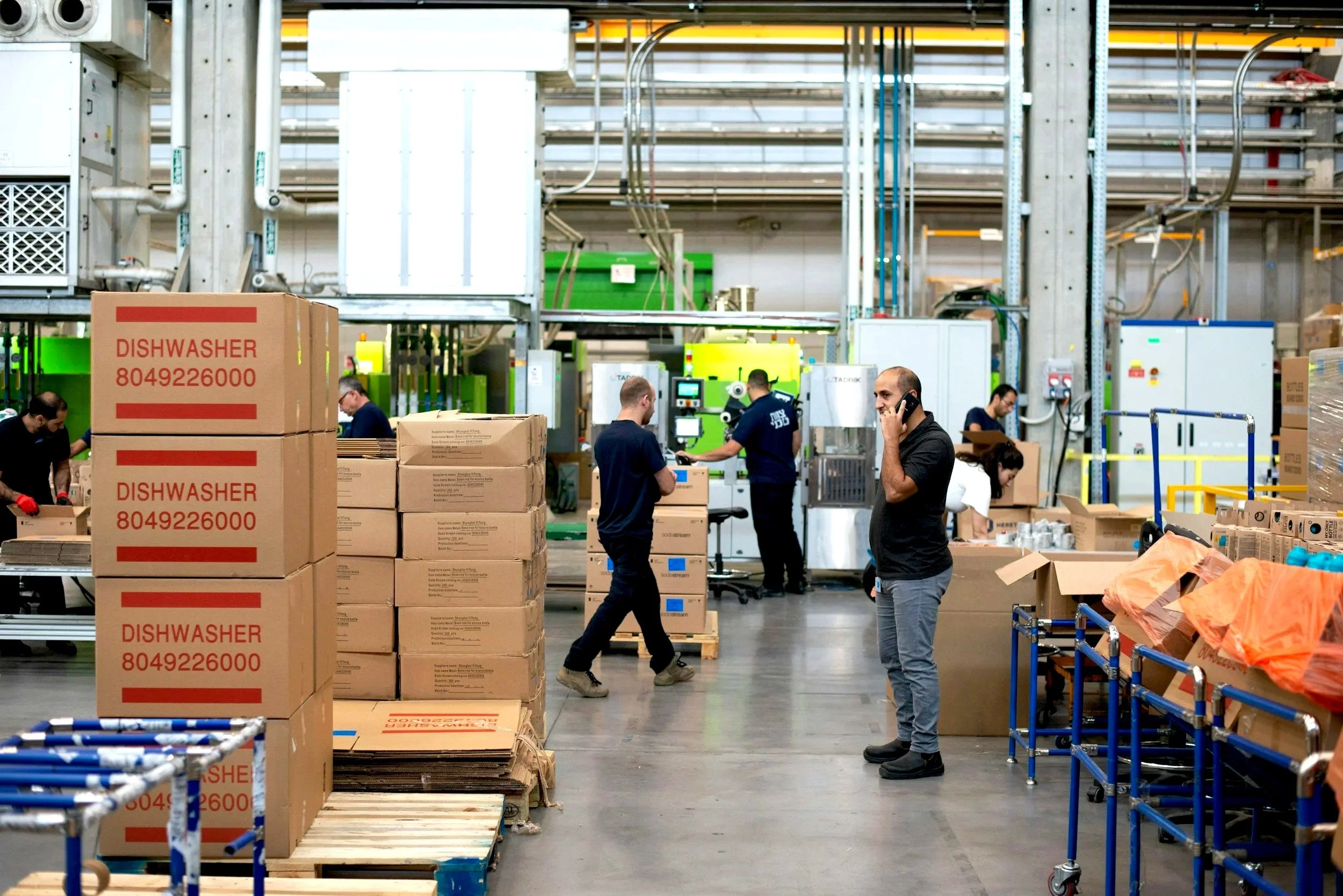 Warehouse interior with workers handling boxes, industrial machinery, and stacks of cardboard boxes, some labeled 'DISHWASHER 804926000'.