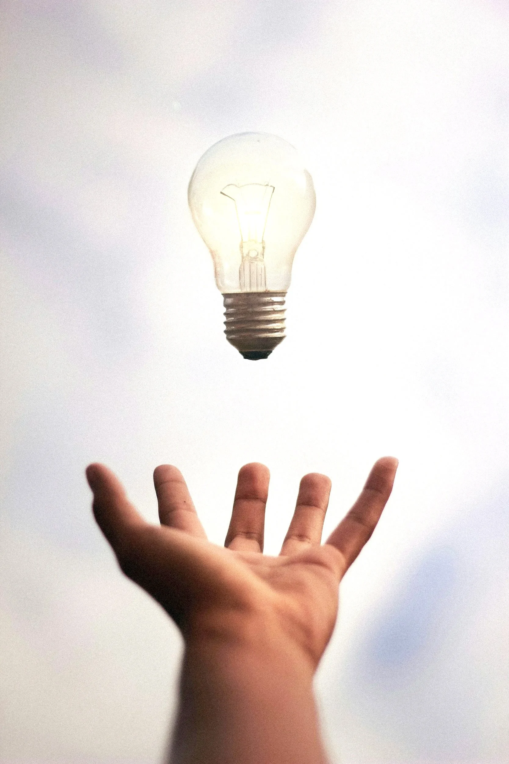 A person’s hand reaching up towards a floating light bulb against a cloudy sky.