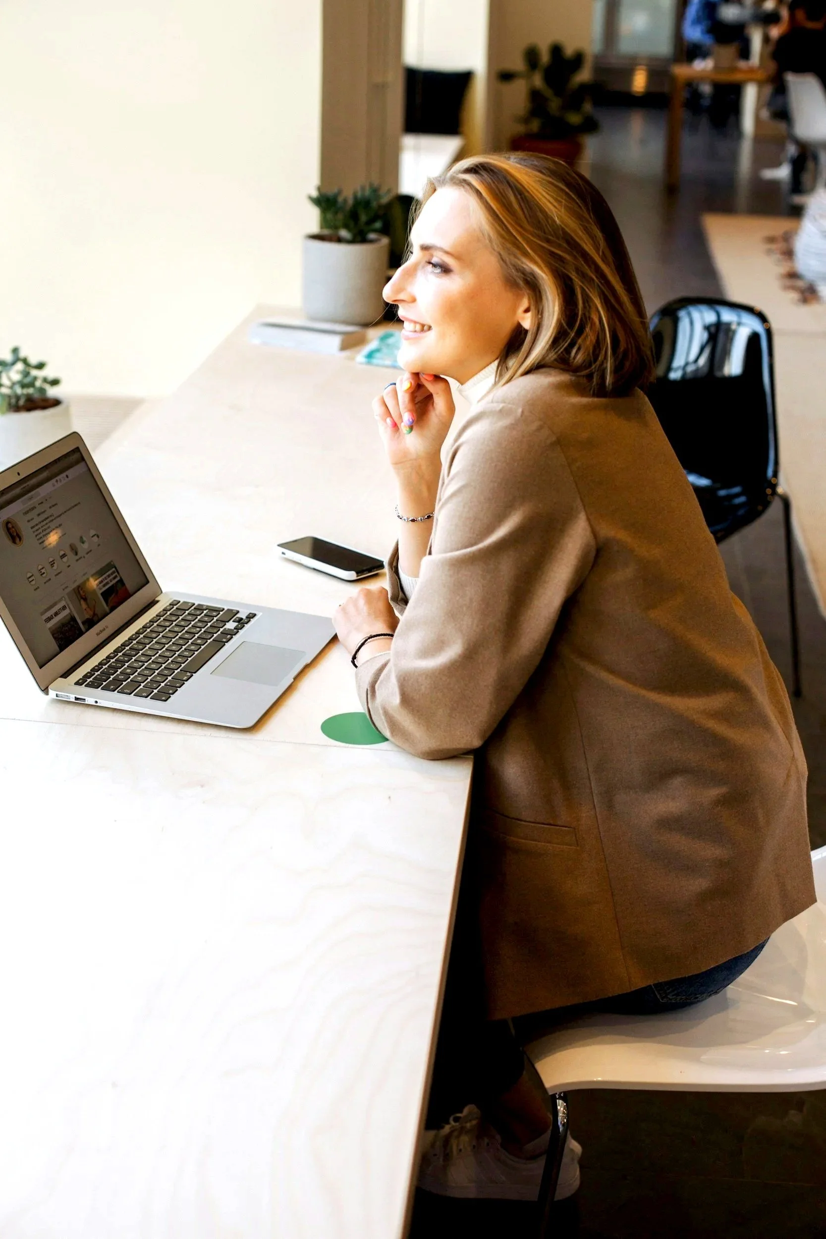 A woman with shoulder-length hair sitting at a light-colored table, looking to the side and smiling, with a laptop, smartphone, and potted plants in the background.
