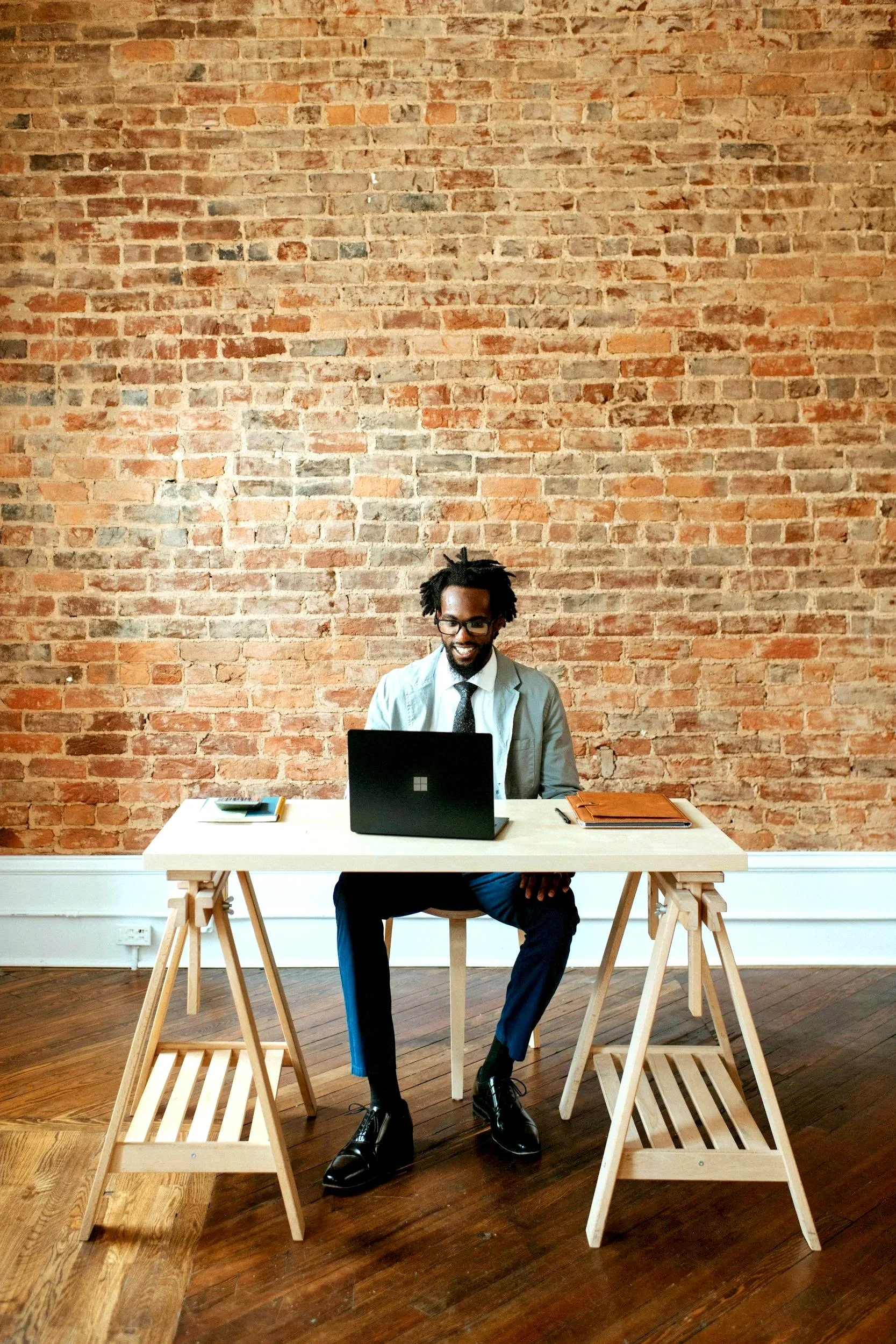 A man with glasses, a tie, and a blazer is sitting at a white desk with a laptop, phone, and folders, smiling in front of a brick wall in a modern office or studio space.