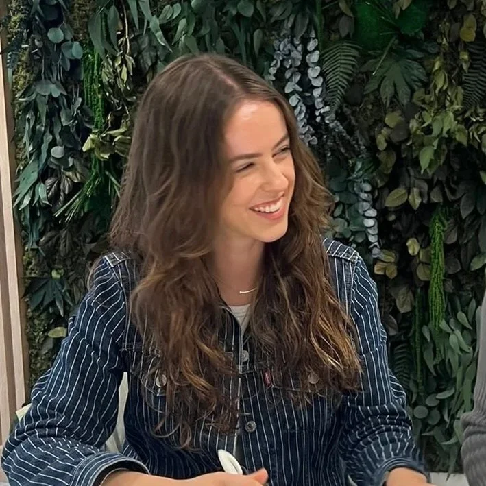 A young woman with long, wavy brown hair smiling and winking, sitting in front of a lush green plant wall.