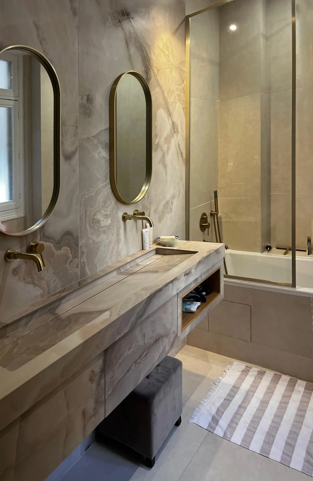 Salle de bain élégante avec un mur en marbre, deux miroirs ovales, un lavabo en marbre, une baignoire avec paravent en verre, et un tapis rayé beige et blanc.